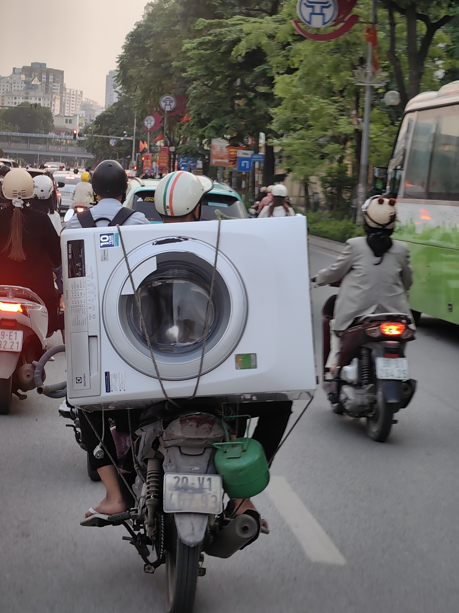 The image captures a bustling street scene in Hà Nội, Vietnam, during what appears to be late afternoon or early evening, characterized by soft, hazy light. The central and most prominent subject is a large white Electrolux front-loading washing machine being transported on the back of a motorbike. The appliance is secured with ropes, indicating a common, makeshift method of goods transport in the region.
The motorbike carrying the washing machine is driven by an individual whose legs and feet, wearing flip-flops, are visible. Another person, likely the driver of a different motorbike, is seen in the foreground with a black helmet and backpack, positioned in front of the washing machine. To the right, another motorbike rider in a light-colored jacket and a beige helmet with a ponytail is seen, with a green and white bus behind them. In the mid-ground, multiple other motorbikes and cars are visible, indicating heavy traffic.
Notable details include the Electrolux brand and text "TIME MANAGER" and "10 YEAR INVERTER MOTOR WARRANTY" clearly visible on the washing machine. The license plate on the motorbike carrying the washing machine reads "20-V1 467.93." A small green container, possibly an auxiliary fuel tank, is attached to the side of the motorbike. Distinctive red and white circular signs with blue symbols, typical street furniture in Vietnam, are visible overhead among the trees. Tall buildings rise in the background, set against a pale sky. The scene illustrates the everyday activity of navigating urban traffic and the resourcefulness in transporting large items within the city.