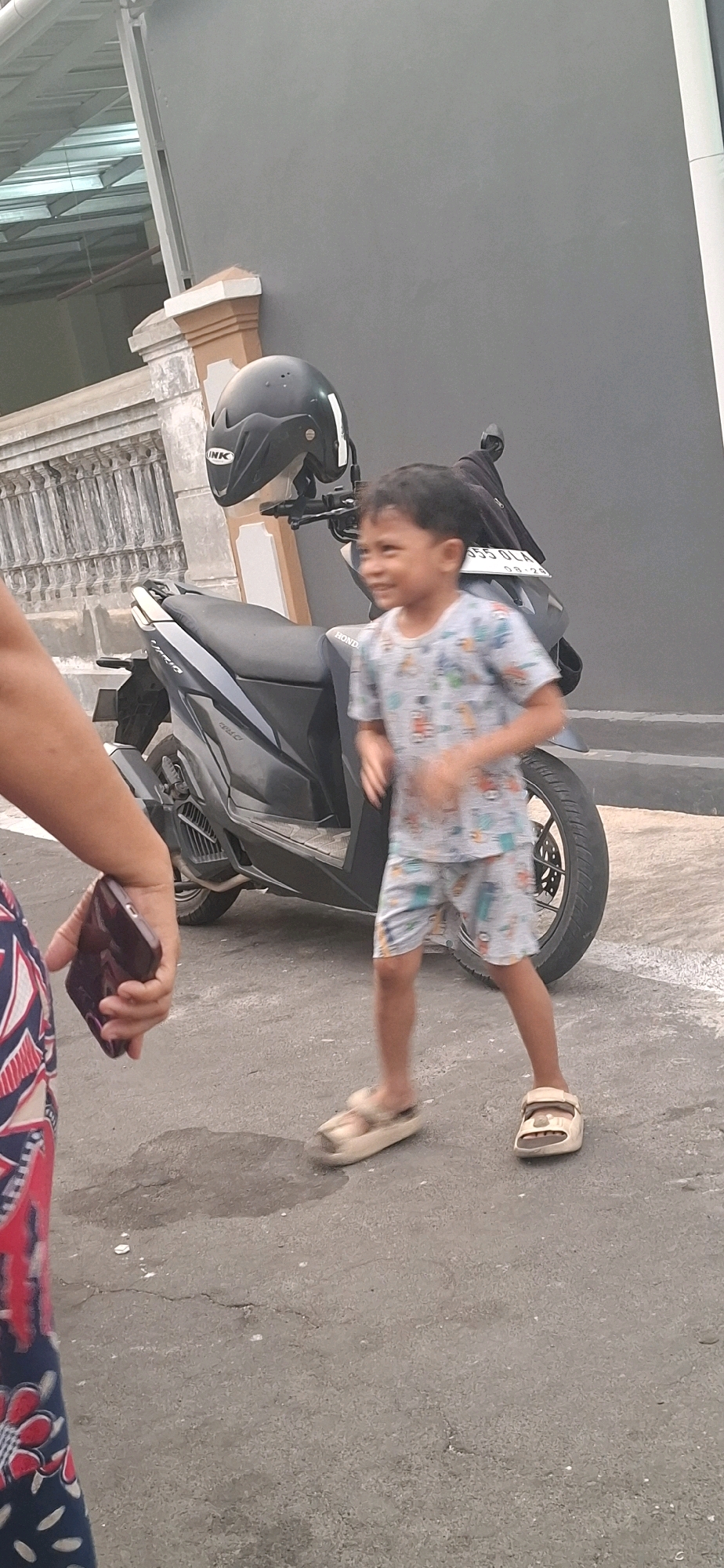 In the foreground, a person's arm and shoulder are visible, holding a dark-colored smartphone. The person is wearing a dark blue garment with a red and white floral pattern. To the right of the frame, a young boy is in motion, his body slightly blurred due to movement. He is wearing a light-colored t-shirt with a multi-colored cartoon print and light-colored shorts with a similar pattern. He is also wearing light-colored sandals.
Behind the boy is a dark-colored scooter, its license plate reading "B 355 OLA" and "03-25". A black helmet with a white "INK" logo is placed on the handlebars of the scooter. The scooter is parked on an asphalt surface, which shows signs of wear and a small wet patch.
In the background, a gray wall of a building is visible. To the left, there is a white concrete railing with decorative elements and a portion of a white roof structure with visible metal beams. The environment appears to be an outdoor residential area, likely in Desa Gagaksipat, Indonesia, given the presence of a scooter and the architectural style. The lighting suggests it is daytime.