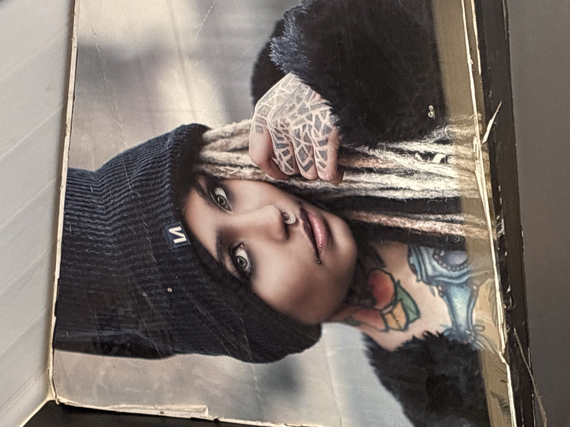 The image is a close-up portrait of a young woman wearing a black beanie and a black fuzzy jacket. She has blonde dreadlocks, septum and nostril piercings, and extensive tattoos on her neck and hands. The background is out of focus and appears to be an urban or industrial setting with muted colors. The image is displayed on a surface that shows signs of wear and tear, with visible scratches, creases, and peeling edges, suggesting it is a photograph or poster that has been handled extensively. The lighting is soft and diffused, highlighting the woman's features and tattoos. The image does not contain any elements that would indicate a specific location in Alanya, Turkey.