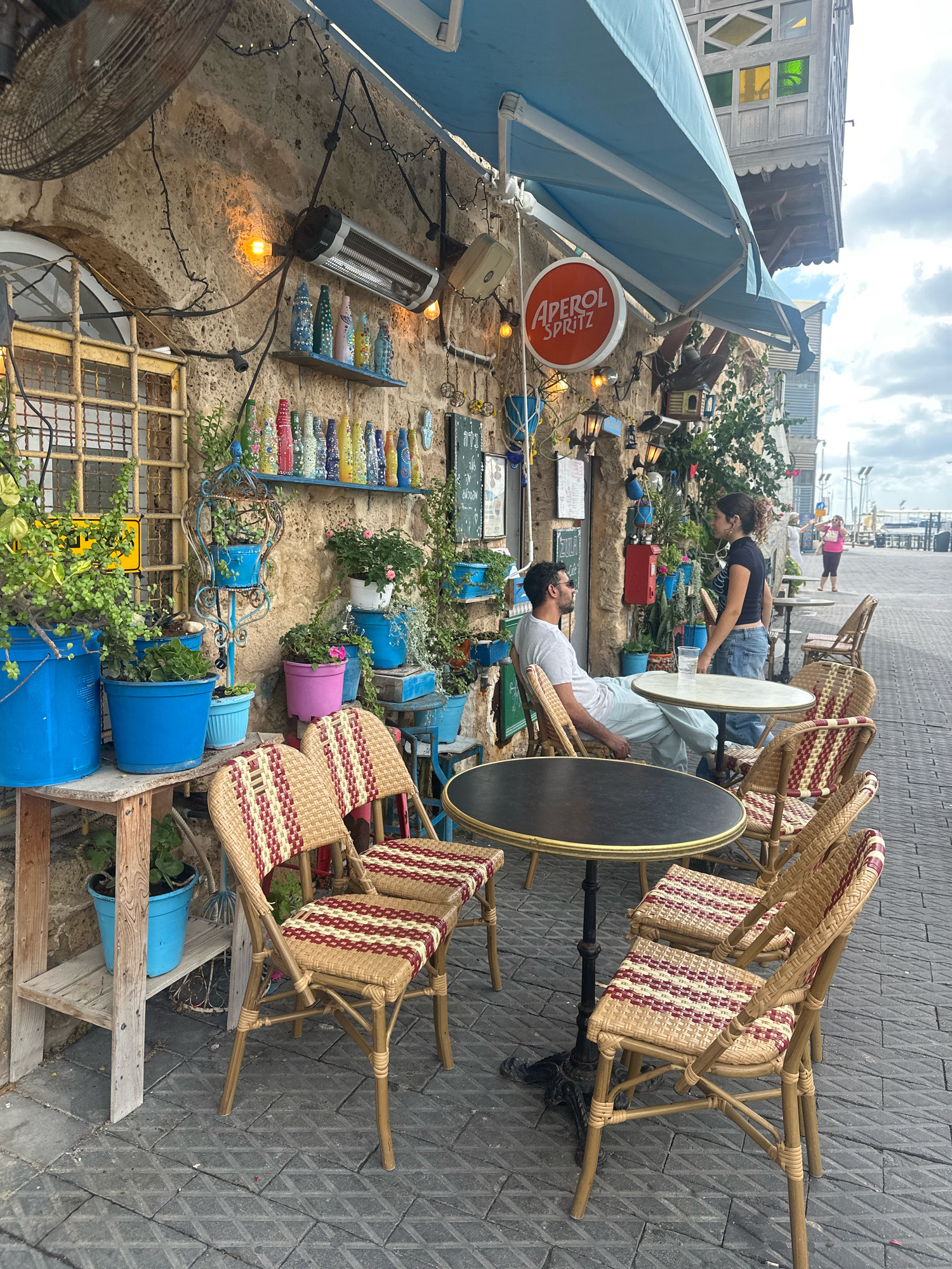 The image depicts an outdoor seating area of a cafe or restaurant located in Jaffa, an ancient port city that is part of Tel Aviv-Yafo, Israel. The cafe's facade is made of rough, stone blocks, and it is adorned with numerous potted plants and decorative items, including a shelf lined with colorful bottles. A blue awning is extended over the seating area, offering shade.
Two people are seated at a small, round table. A man wearing a white t-shirt and light-colored pants sits facing left, while a woman in a black t-shirt and blue jeans stands, facing the man and gesturing. A glass of water is on the table between them.
The seating area features several chairs made of rattan or wicker with a red and beige striped cushion. Another table is visible in the foreground. The ground is paved with square tiles arranged in a diamond pattern. In the background, a promenade and the sea can be seen, with boats docked and people walking. The sky is partly cloudy. The overall scene suggests a relaxed, bohemian atmosphere typical of certain areas in Jaffa.