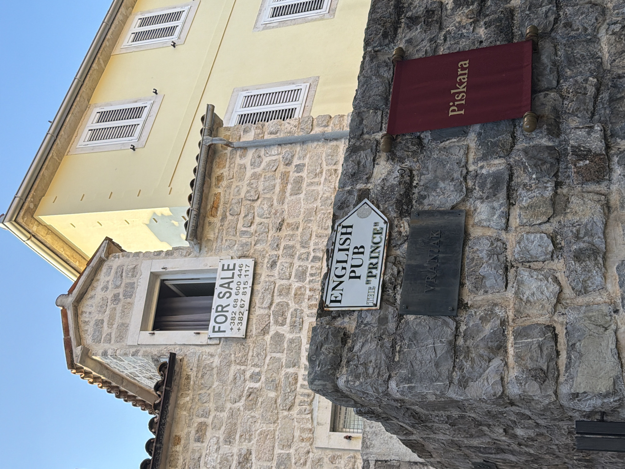 This is a photograph taken in Budva, Montenegro. The image shows a stone wall in the foreground with several signs attached. A sign for an "ENGLISH PUB THE 'PRINCE'" is pointing to the right. Below it, a dark plaque reads "VRANJAK". To the right of these signs, a maroon banner with the word "Piskara" in gold lettering is hanging. Behind the stone wall, a building constructed with light-colored stone has a "FOR SALE" sign with phone numbers displayed in a window. Further in the background, a pale yellow building with white shutters is visible under a clear blue sky. The sunlight suggests it is daytime.