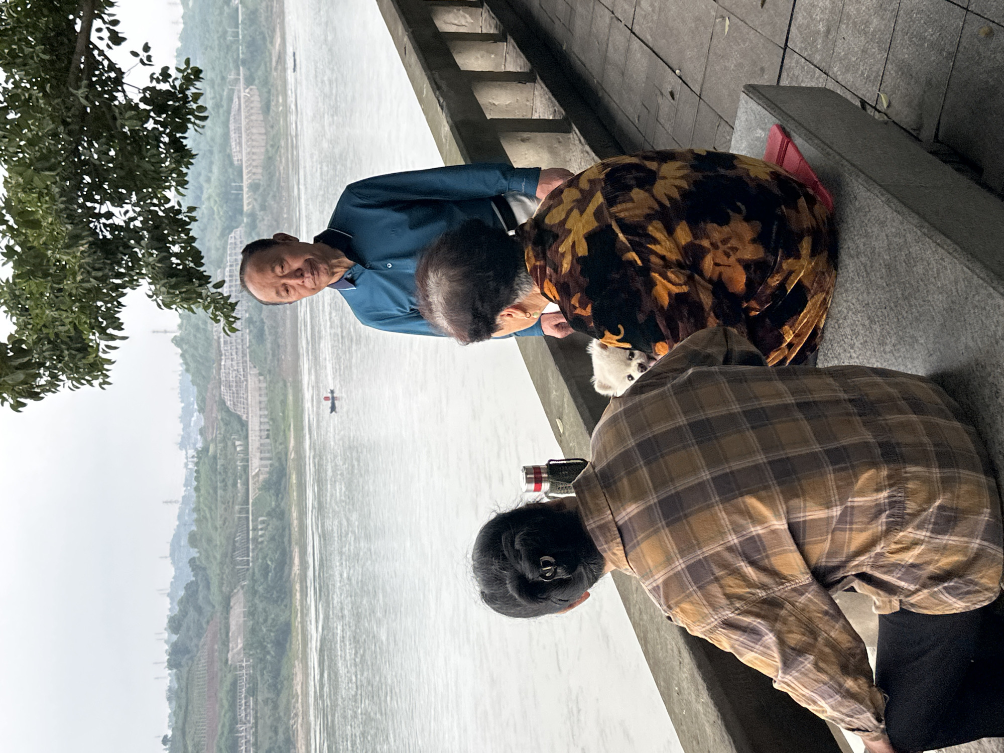 Three individuals and a dog are gathered alongside a riverbank in Jiangjin, China. An elderly man in a blue shirt is visible to the left, while two other individuals, one wearing a floral patterned garment and the other a plaid shirt, are positioned closer to the river's edge. A small white dog is situated near the floral garment. A water bottle rests near the person in the plaid shirt. A waterway stretches into the distance, with visible structures and land along the opposite bank. Foliage overhangs the scene from the upper-left portion of the image.