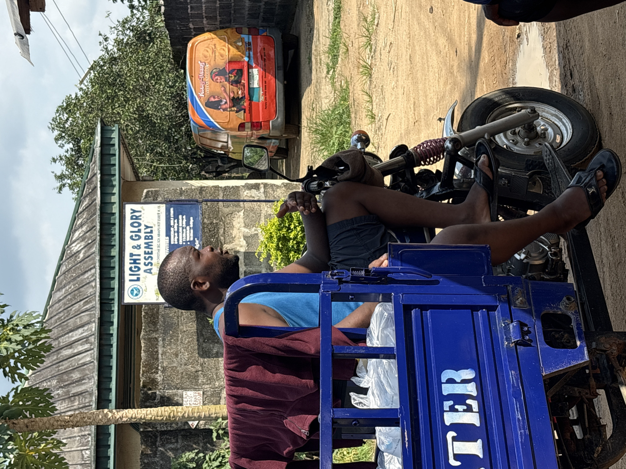 A man is on a blue three-wheeled vehicle in Ikot Ekpene, Nigeria. The vehicle has “TER” printed on its side. The man wears a blue tank top and gray shorts, and he is sitting in the vehicle. In the background is a building with a sign for “LIGHT & GLORY ASSEMBLY,” which says it is “BY CHRISTIAN SEC, SOL ABIAKPO KOT ESSIER IKEL.” A maroon towel hangs on the side of the vehicle. A car with the inscription “TUMMY TOMM INSTANT NOODLES” is parked nearby. The environment includes trees, grass, and dirt.