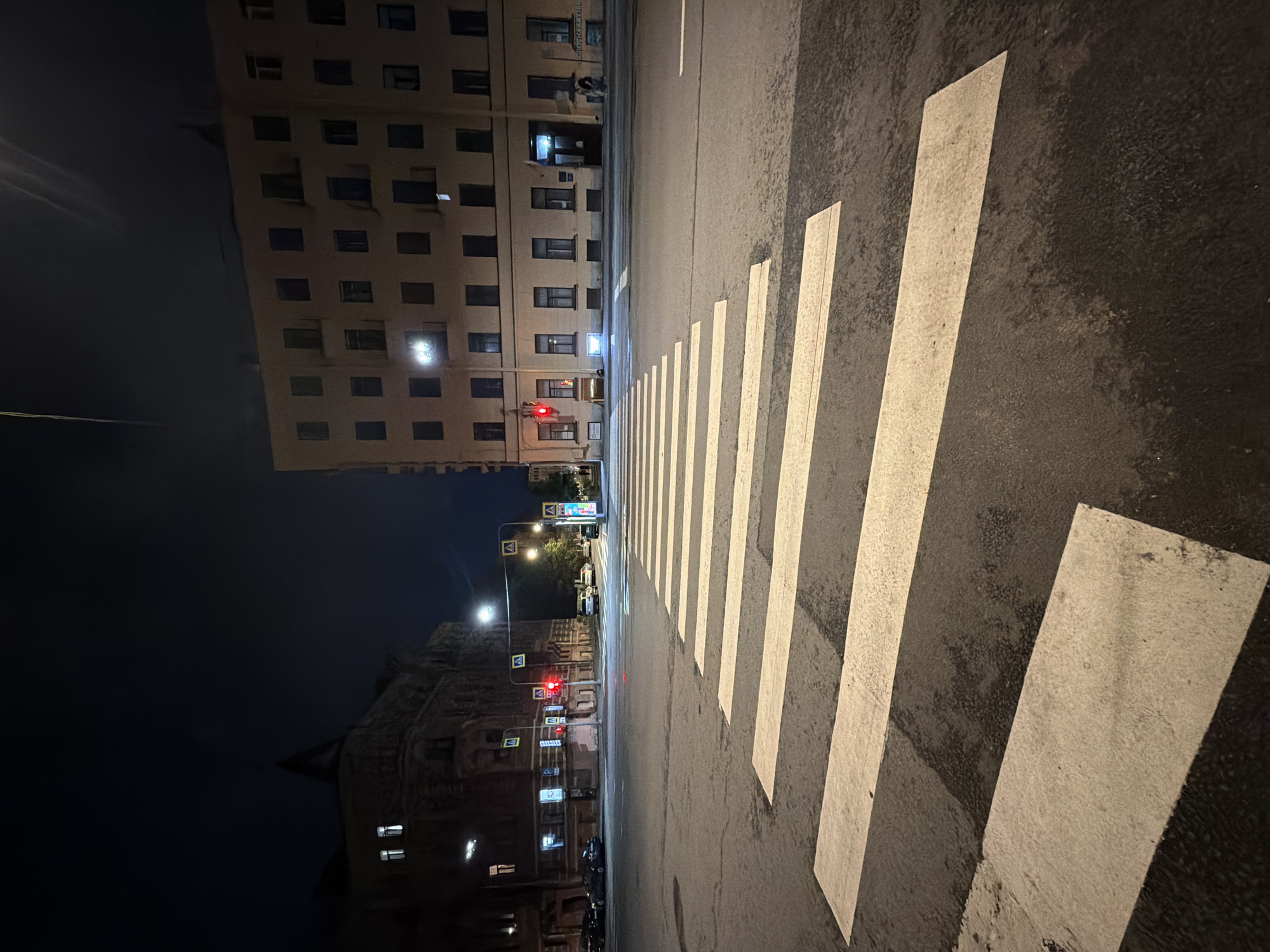 The image captures a street scene at night in Saint Petersburg, Russia, likely a crossroads with a prominent pedestrian crossing. The perspective is low, emphasizing the white stripes of the crosswalk in the foreground. On the left, a dark, older building with illuminated windows and ornate architectural details stands against the night sky. To the right, a lighter-colored, multi-story building with numerous dark windows dominates the frame. Traffic lights are visible, one displaying a red light. Several cars are present on the street, and streetlights cast illumination on the road and buildings. The overall atmosphere is quiet and urban, typical of a city at night.
