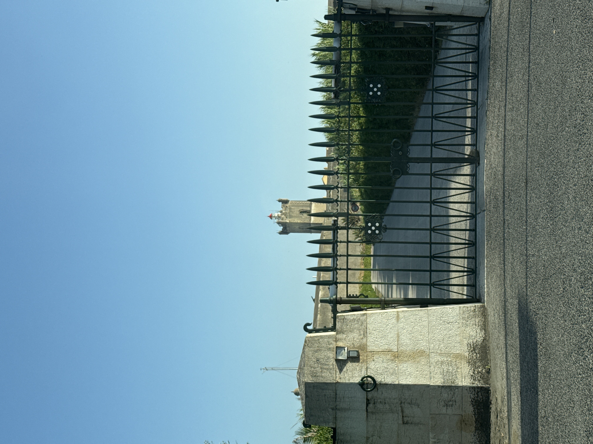An imposing dark green metal gate with spiked finials stands closed in the foreground, obscuring a paved driveway. The gate is flanked by large, rough-hewn stone pillars. The pillar on the left has a circular metal ring attached and a small square electrical box mounted on its surface. Beyond the gate, the driveway leads towards a historic stone tower with a red-capped lantern room, identified as the São Lourenço Lighthouse, located in the city of Oeiras, Portugal. The lighthouse is situated within a walled complex, bordered by dense green foliage. The sky above is a clear, bright blue.