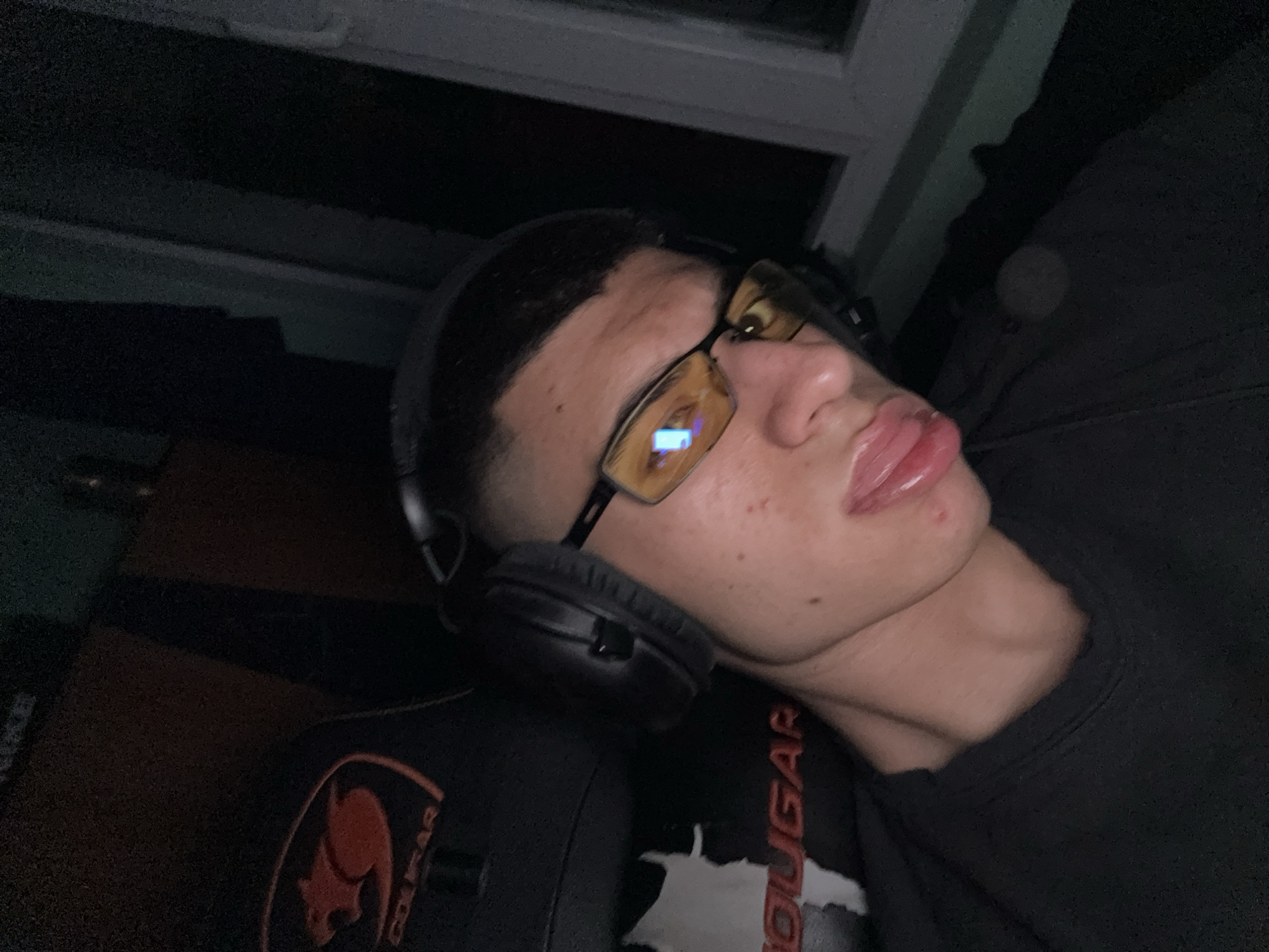 A medium, high-angle indoor shot shows a male wearing black eyeglasses with yellow-tinted lenses and black headphones lying against a black gaming chair branded "COUGAR." The male is wearing a black shirt. Above the subject is a window. The location is not discernible from this image.