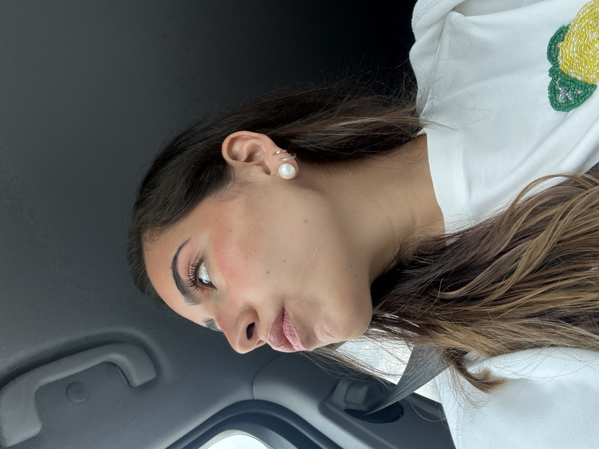 A woman with long, brown hair and pearl earrings is shown in profile. She is wearing a white t-shirt with a lemon design and is buckled into a car. The interior of the car is visible, with a gray roof and grab handle. The location cannot be determined as San Antonio because there are no objects or landmarks that are clearly visible in the photo.