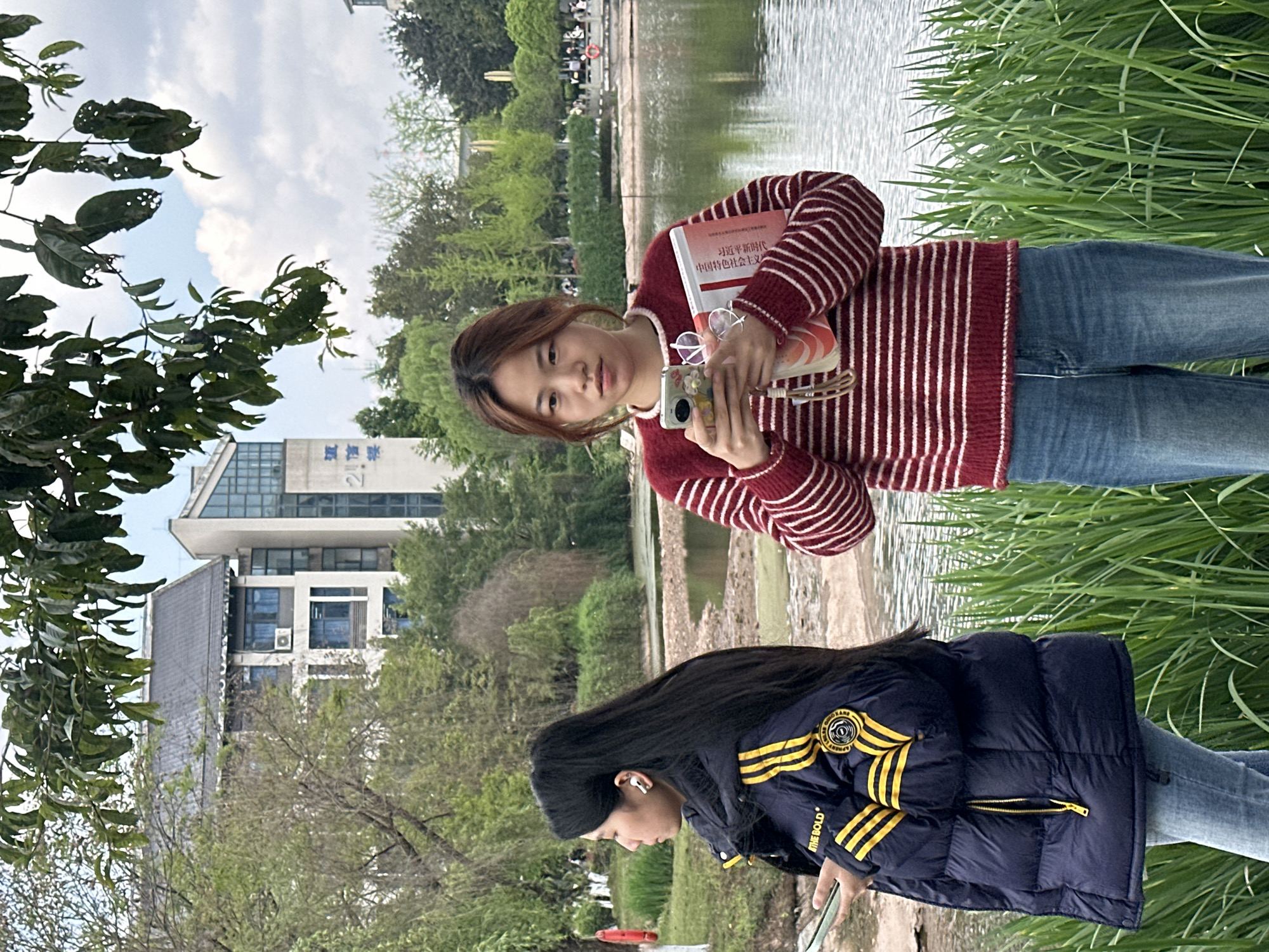 Two women stand near a body of water on a sunny day in Jiangjin, China. One woman, wearing a red and white striped sweater and jeans, holds a book and a camera. The other woman, wearing a dark blue puffer jacket with yellow stripes and jeans, looks down towards the ground. In the background are buildings, trees, and a lake. The location appears to be a park or campus setting.