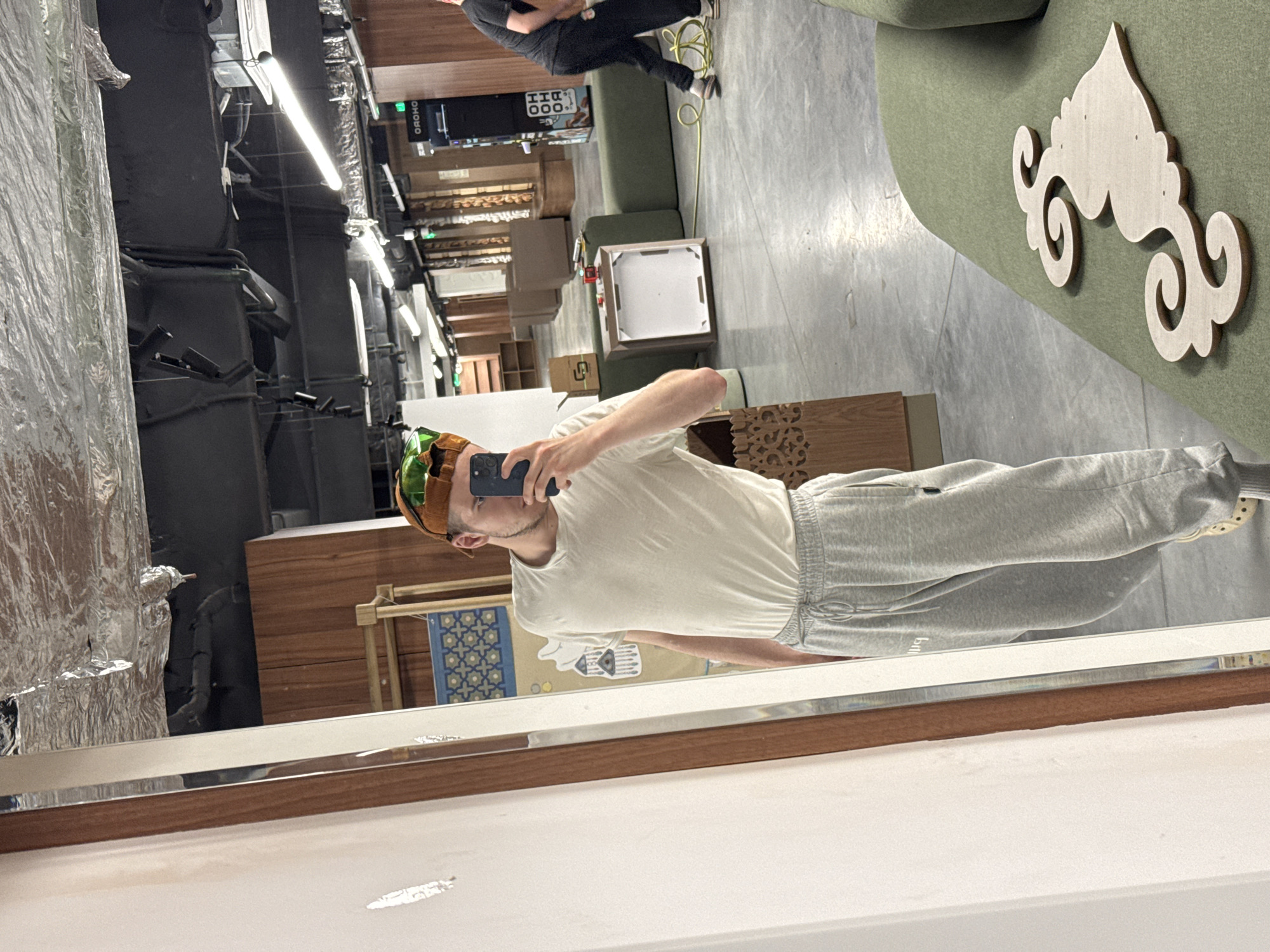 A young man with light brown hair and a tan complexion is taking a mirror selfie. He is wearing a white t-shirt and grey sweatpants. On his head, he is wearing a novelty hat that resembles a hamburger. He is standing in a room that appears to be a workshop or studio. In the background, there are shelves, furniture, and a vending machine. There is also another person visible in the background, wearing a black t-shirt and black pants. The floor is concrete and there is a large mirror in the foreground. The overall environment is industrial.