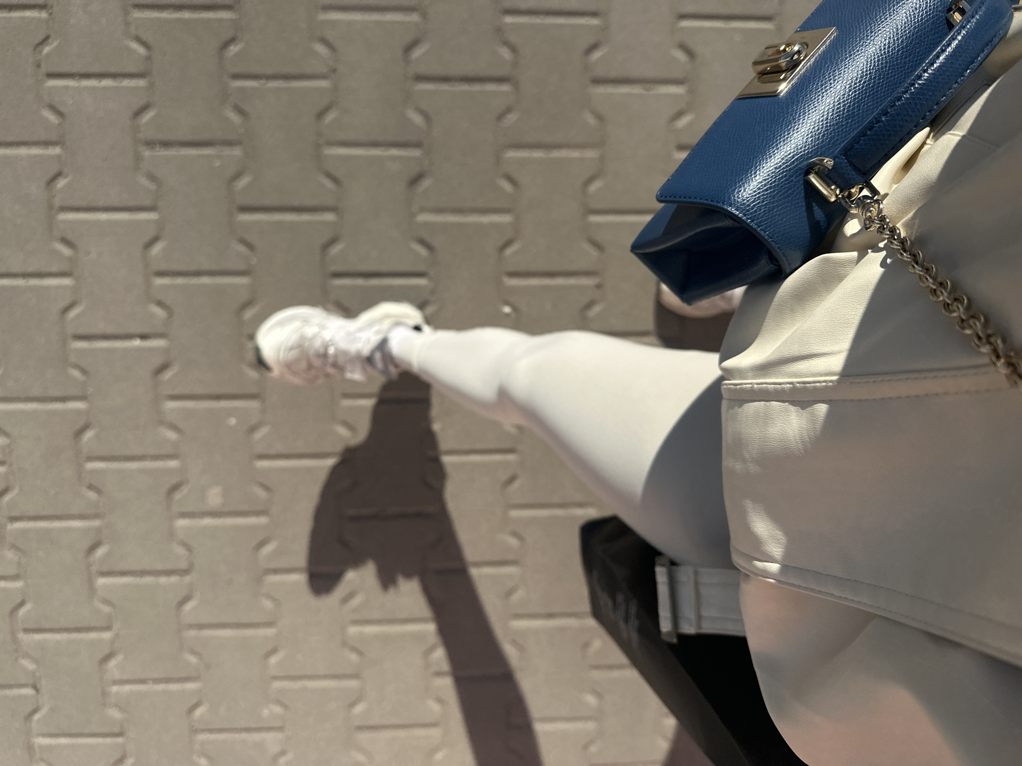 A waist-down, high-angle shot depicts a person standing on a grey brick sidewalk. The sidewalk bricks are arranged in a repeating pattern. The person wears white leggings and white sneakers, with a shadow stretching diagonally across the sidewalk. A white jacket and a small blue purse with a silver chain are also visible on the right side of the frame. The location is likely a street in Sofia, Bulgaria, given the type of brick used on the sidewalk which is a common feature in many cities in Bulgaria and especially in Sofia.