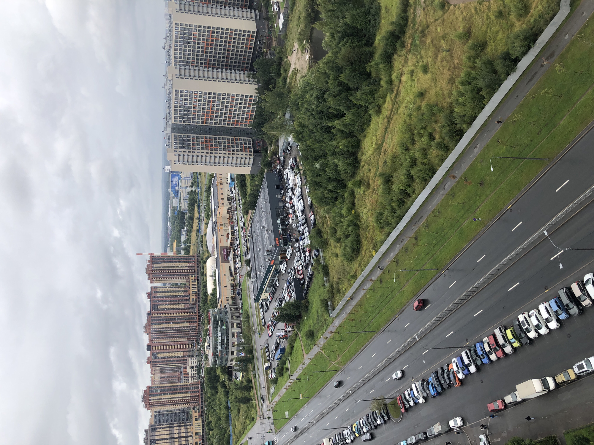 An aerial view captures a cloudy day in a city, featuring several large, multi-story residential buildings with beige and reddish-brown facades. In the foreground, a multi-lane road runs diagonally, wet from recent rain, with various cars parked along its edges and in designated parking areas. To the right of the road, a strip of green grass is bordered by a white fence. Further to the right, a dense cluster of green trees surrounds a small body of water, and beyond that, more residential buildings are visible. A low-rise building with a circular glass facade is situated between the road and the taller buildings. Several cars are parked in front of a long, low-rise building that appears to be a car dealership, identifiable by the "LADA" signage. A few individuals are seen walking on the sidewalk next to the road. The sky is overcast with gray clouds.