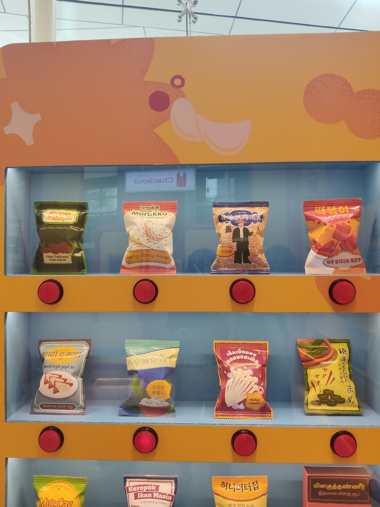 The image shows a vending machine displaying various snacks. The vending machine is positioned indoors, likely in a public space such as a mall or transit hub, given the presence of a "CHECKOUT" sign in the background. The machine has multiple shelves with individually packaged snacks, each featuring distinct graphics and text in different languages, suggesting an international product assortment. Each item is presented with a red circular button beneath it, presumably for selection. The overall color scheme of the vending machine is bright yellow and orange, with a light blue interior for the display areas. The snacks on display appear to be chips, crackers, or similar savory snacks. The snacks are arranged in two rows, with four items on the top shelf and four on the middle shelf visible. The bottom shelf is partially visible, showing at least two more items. There are no people visible in the frame.