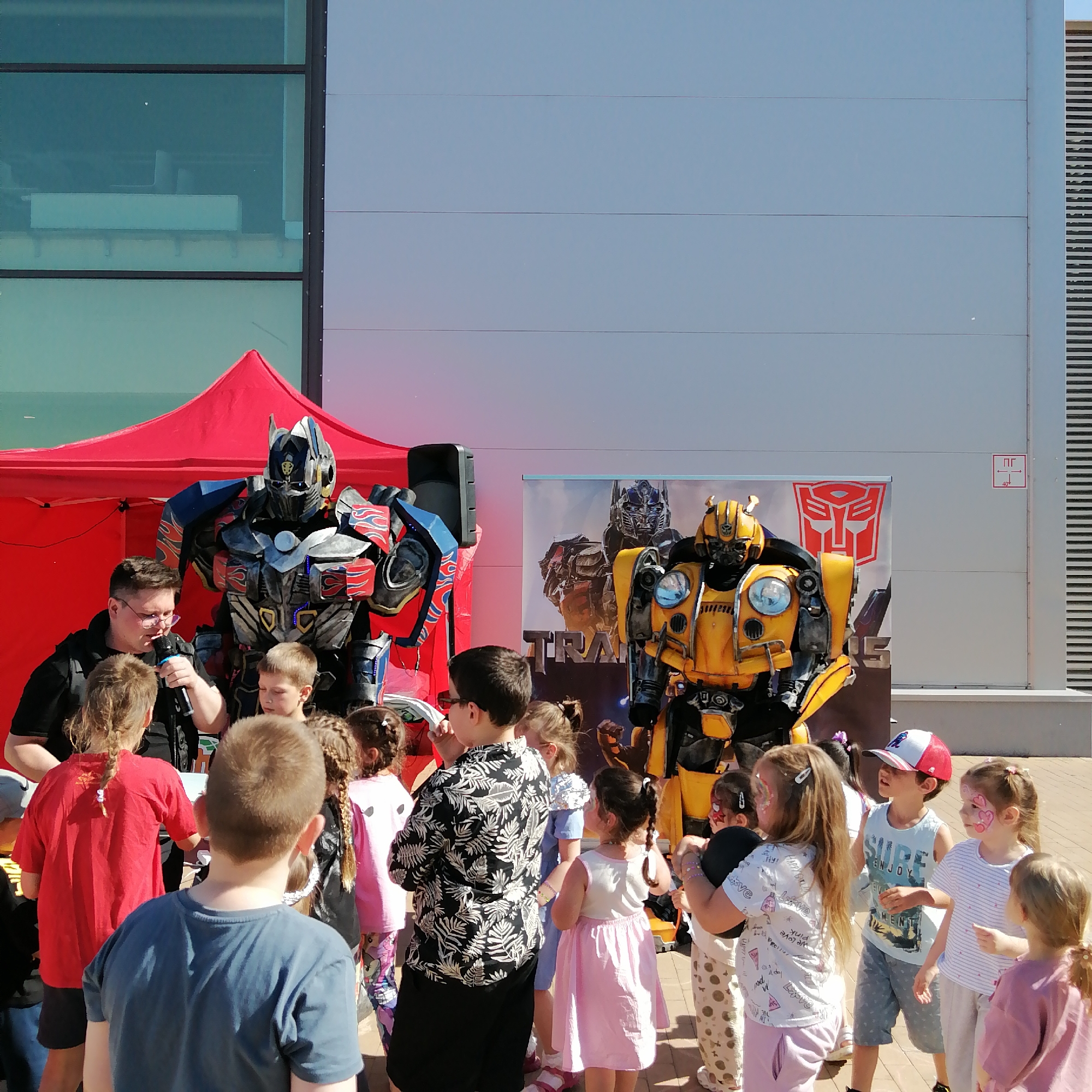 An outdoor scene in Трубачеевка, Russia, features two individuals in detailed Transformers costumes, Optimus Prime and Bumblebee, surrounded by a group of approximately 10-15 children. The Optimus Prime character, in red, blue, and grey armor, stands beneath a red canopy tent on the left. Adjacent to Optimus Prime, an adult male wearing a dark shirt and glasses holds a microphone and interacts with the children. A black speaker on a stand is positioned behind him. The Bumblebee character, in yellow and black armor, stands further to the right, in front of a large banner displaying images of Transformers characters and the Autobot insignia.
The children, appearing to be of elementary school age, are gathered in the foreground and midground, intently observing the characters. Several children have face paint, including a girl with a heart and star design and another with a spider-like motif. Notable children include a boy in a blue t-shirt facing away from the camera in the immediate foreground, a girl in a red t-shirt with braided hair facing away near the speaker, and a boy in a white and red baseball cap and "SURF ENJOY" tank top looking up at Bumblebee. A girl in a light pink dress stands near Bumblebee. Another adult male, wearing a black and white patterned shirt, is partially visible among the children on the right.
The background consists of a modern building with large, reflective glass windows on the left and light grey or blue-grey horizontal paneling covering the majority of the wall. A red and white utility sign marked "ПГ 40°" is visible on the building's paneling on the right. The ground is paved with light-colored interlocking pavers. The scene is illuminated by bright daylight, suggesting a clear, sunny day.
