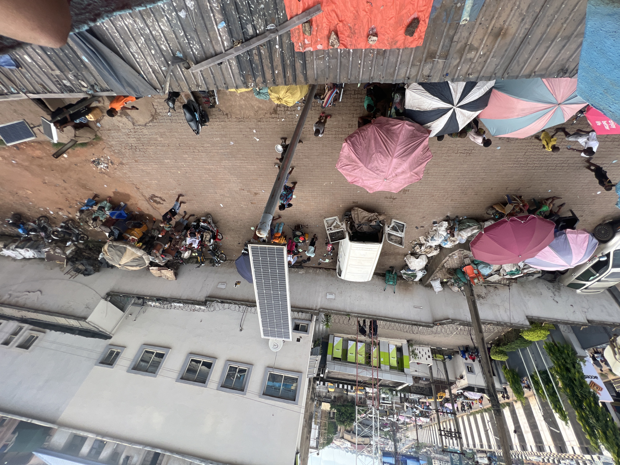 This image provides an overhead view of a bustling street scene in Nkpor, Nigeria, likely during daylight hours given the shadows. The setting appears to be a marketplace or a busy commercial area with a mix of structures, including buildings with windows and awnings, and open ground paved with interlocking bricks.
Several people are visible throughout the scene, engaged in various activities. Some are walking, others are gathered in small groups, and a few are interacting with objects. Objects include parked motorcycles and bicycles, a white van, and numerous umbrellas providing shade. A large solar panel is mounted on the side of a building, suggesting an effort towards renewable energy. There are also piles of what appear to be goods or waste materials.
The presence of multiple umbrellas, some colorful and others plain, indicates a sunny day, and they are used by people to shield themselves from the sun. The overall atmosphere seems active and lively, typical of a busy public space in a Nigerian town.
There is some visible text. One sign on a building reads "ATM" in large green lettering. Another sign, partially visible and angled, appears to have the word "ACCE" on it. The overall impression is a snapshot of everyday life and commerce in Nkpor.