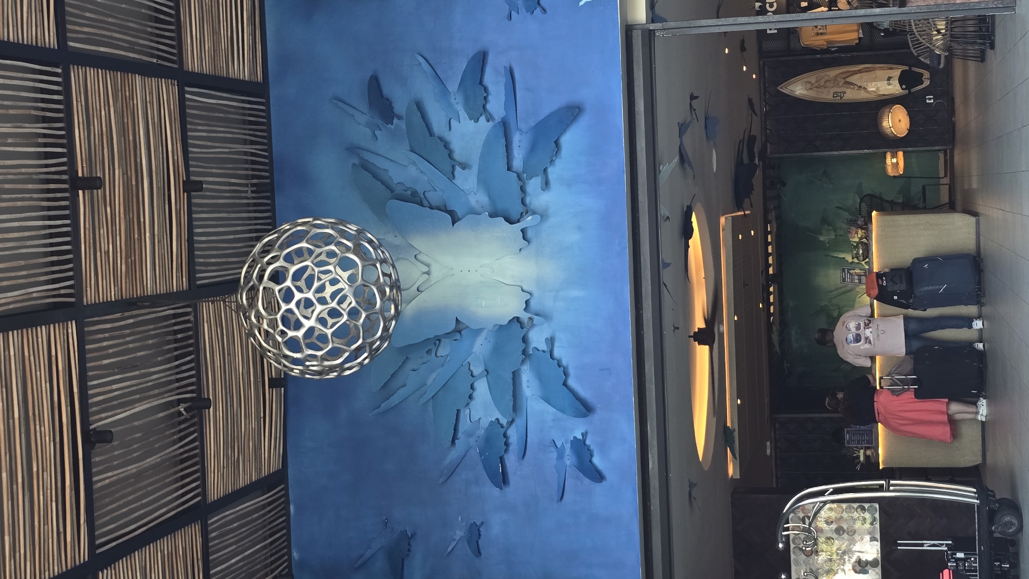 The image captures a scene in what appears to be a hotel or resort lobby, likely in Kuta, Indonesia, given the architectural elements and decorative style. The lobby features a striking blue wall adorned with a butterfly-themed art installation. A spherical, patterned light fixture hangs adjacent to the wall.
On the right side of the frame, a partial view of the lobby reveals a reception area with a counter. Behind the counter, there's artwork on the wall. Two individuals are present; one wearing a white graphic T-shirt and jeans, and another wearing a dark top and a pink or red skirt. They are either checking in or out, as luggage is near them. A porter's trolley is positioned near the reception area.
Above the reception, a surfboard is mounted on the wall along with lighting fixtures. The architectural style incorporates a mix of natural and contemporary elements, including what appears to be a bamboo-patterned ceiling or wall feature. The setting suggests a relaxed, tropical environment.