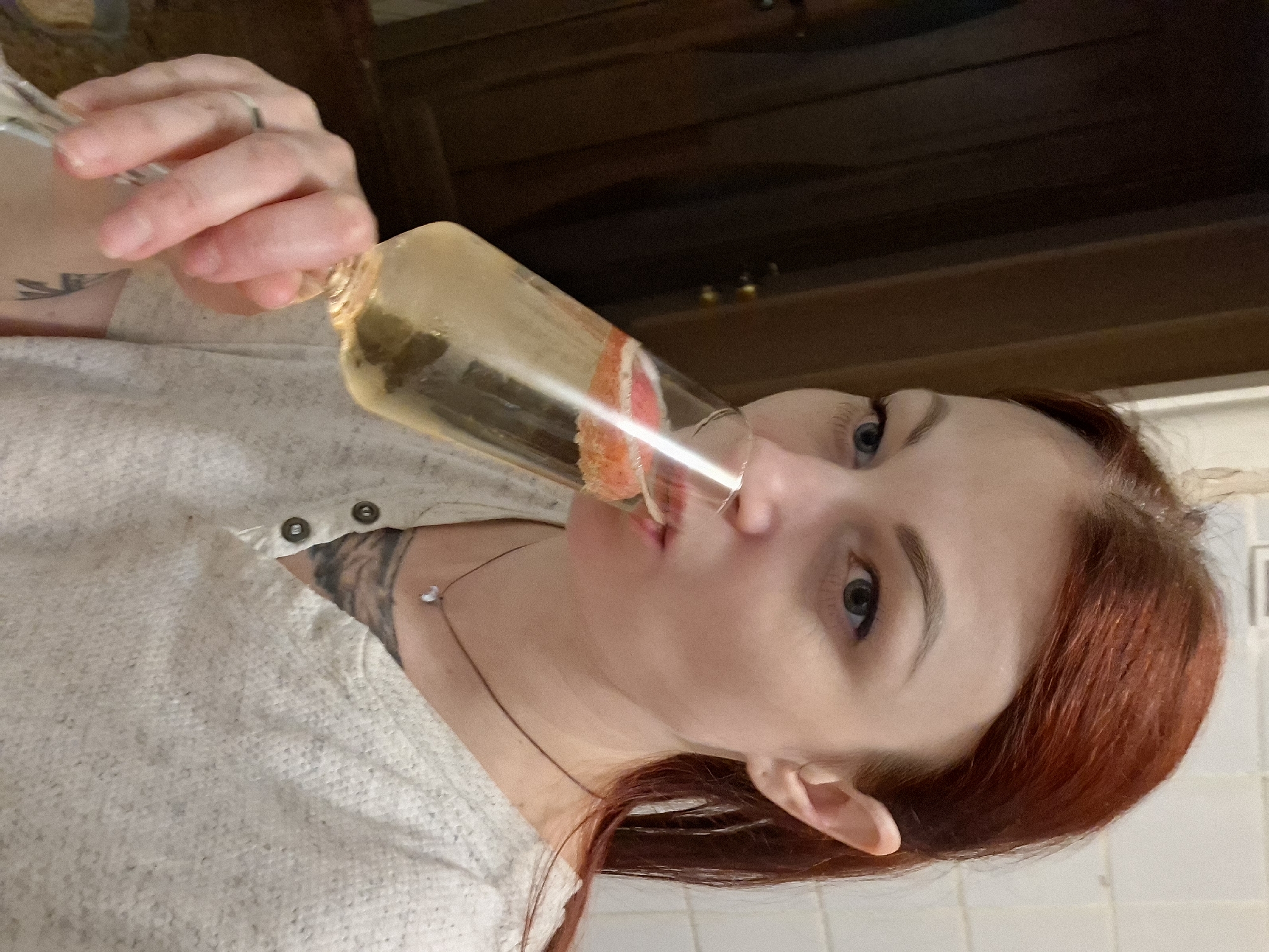 An adult female, appearing in her late 20s to early 40s, with reddish-brown hair, is depicted indoors in Забалоцкі сельскі Савет, Belarus. She is seen in a three-quarter profile, bringing a clear champagne flute to her lips and drinking from it. Her eyes are light-colored (blue/grey) and directed slightly upwards and to the left. She wears a light grey or off-white textured long-sleeved top with a Henley-style neckline featuring two dark buttons. A thin dark cord necklace with a small, light-colored pendant is visible around her neck. Tattoos are partially visible on her left forearm/bicep area and on her chest beneath the shirt neckline. A silver-toned ring is on her left hand's ring finger, which is holding the stem of the glass. The glass contains a light yellow, effervescent liquid, with two slices of red fruit (possibly strawberry or grapefruit) floating within it.
The background on the left consists of a dark brown, vertically paneled surface, possibly a door or furniture. The background on the right features a light-colored tiled wall with visible grout lines. The lighting appears to be internal ambient light, casting subtle shadows on the subject's face and beneath her chin.