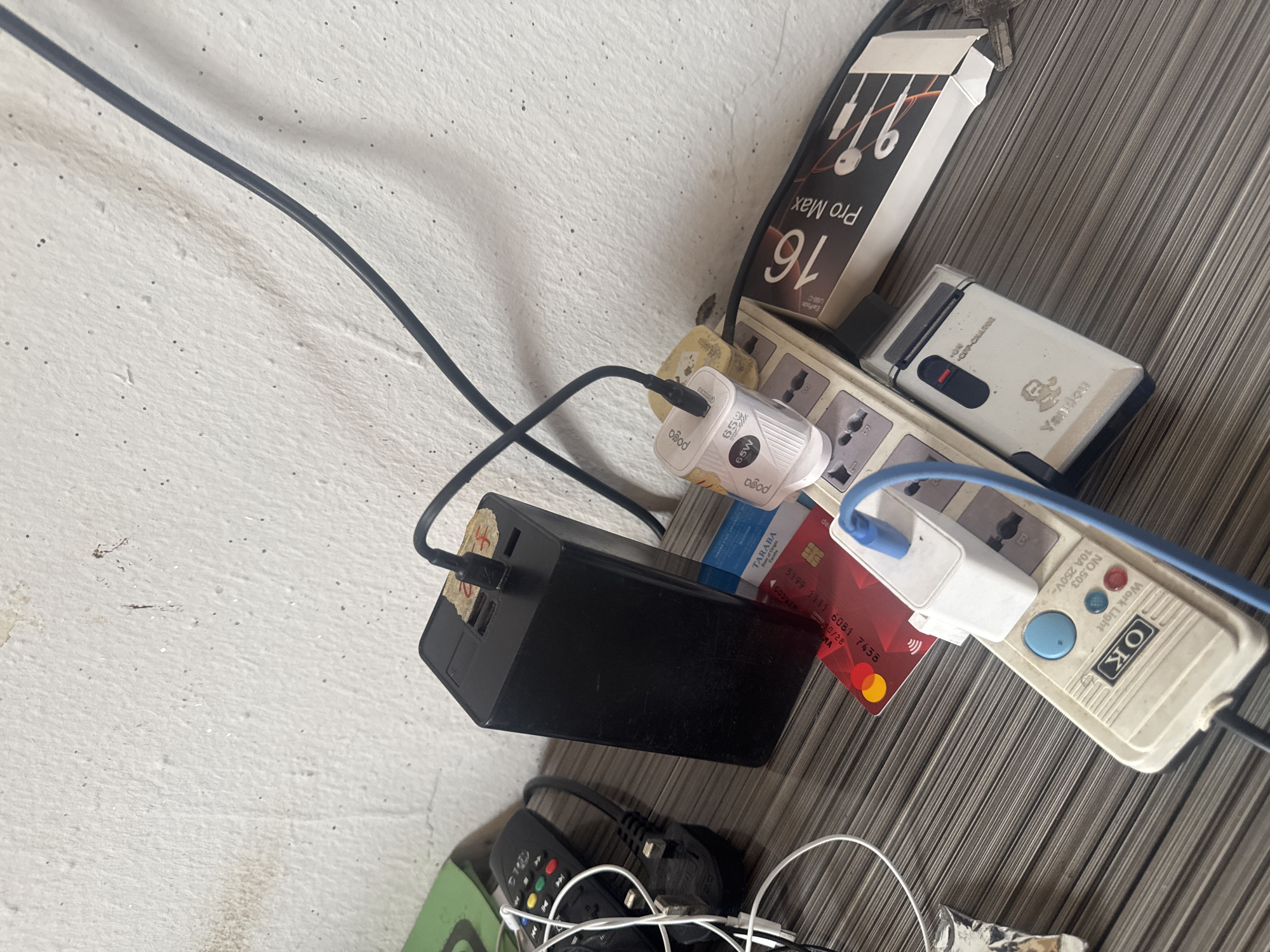 The image depicts a close-up, high-angle shot of a cluttered surface, possibly a desk or shelf, against a textured white wall. The surface is a striped, dark gray material.
Dominating the scene is a large black power bank, with a black USB cable plugged into its top. To the right of the power bank is a white power strip with multiple outlets. Several items are plugged into the power strip: a white charger with "pogo" and "65W" printed on it, and a blue cable connected to another white adapter. A red credit card is also visible beneath the white charger.
Further to the right of the power strip, a box for "Pro Max" earbuds is present. Below the power strip, a silver electronic device with "Yandun" and "ON/OFF-CHARGE" printed on it is visible. To the left of the power bank, a black remote control with colorful buttons and several white cables are present. Some of the cables appear to be connected to a black plug. A crumpled piece of silver foil is also visible in the lower left corner.
The wall behind the objects shows signs of wear, with some discoloration and small dark specks. The lighting is somewhat harsh, casting shadows. The overall impression is one of a functional but disorganized charging station.