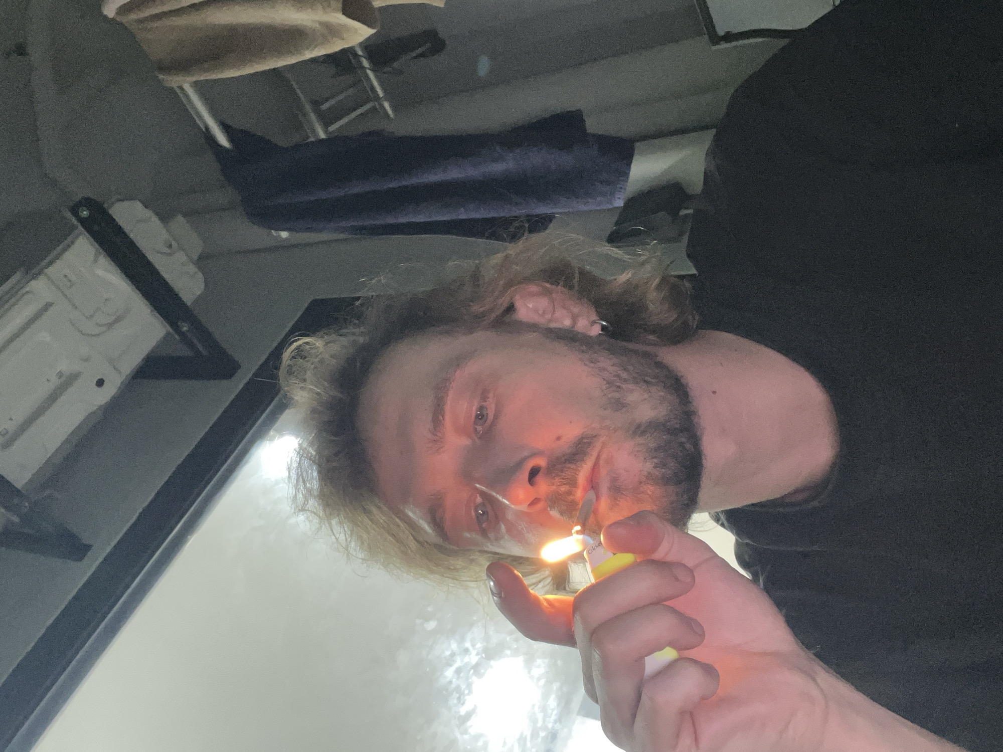 An upside-down close-up photograph captures a man with messy light-colored hair and a beard, looking directly at the viewer. He holds a yellow "Clipper" brand lighter in his right hand, its flame brightly illuminating his face, particularly around his mouth. He appears to be lighting an object that is obscured from view by his hand and the flame. A small earring is visible in his left ear, and he is wearing a dark-colored shirt.
The background is an indistinct indoor setting, featuring what appears to be a light-colored object (possibly part of a bed or shelf) and a folded dark blue fabric. The lighting is low, with the lighter's flame providing a prominent light source, complemented by a bright reflection or light source in the upper right. The image is taken from a low angle, creating a unique perspective. The man's expression is focused and engaging. The photo is provided with the location context of Nam Natural Heritage Area Trang, Vietnam.