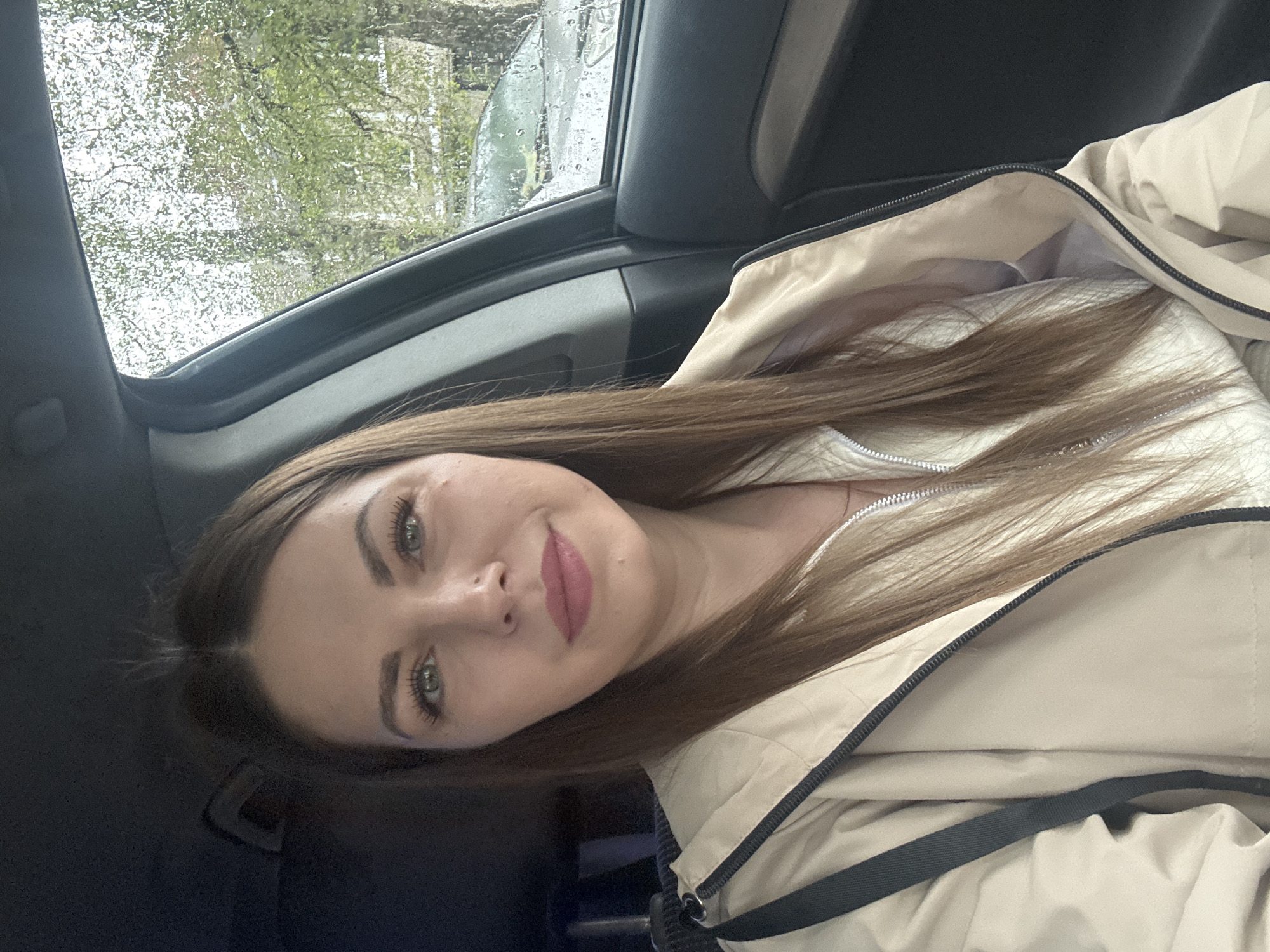 A young woman with long brown hair, green eyes, and pink lipstick is sitting inside a car. The car's window is visible, showing a rainy day outside with trees and another vehicle visible. The woman is wearing a light beige jacket with a black zipper. The interior of the car is dark, with elements like the car door and ceiling visible. While the woman is likely in a car, the image does not contain the location in the city of Раздолье, Russia.