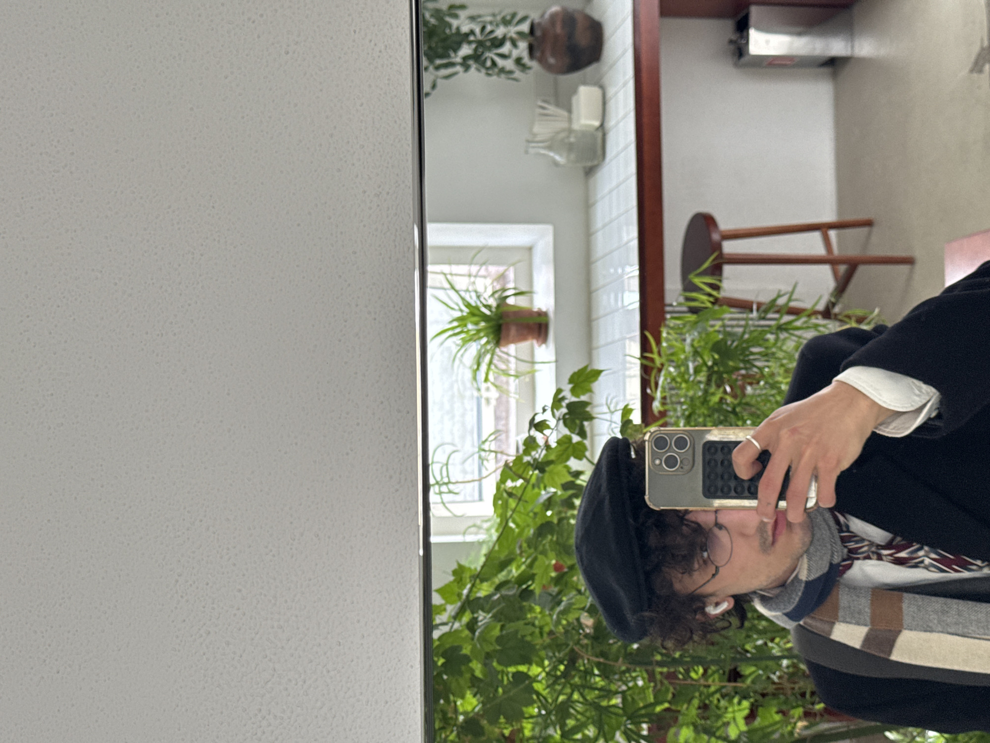 The image shows a reflection in a mirror. The reflection includes a person holding a cell phone up to take a picture, a room interior with plants, and a window. The person is wearing a beret, glasses, a scarf, and a dark jacket with a white cuff showing. The room has a plant on a windowsill, a wooden stool, and other plants in the foreground. There's also a white tiled area. There is no information to determine if this photo was taken in Moscow, Russia.