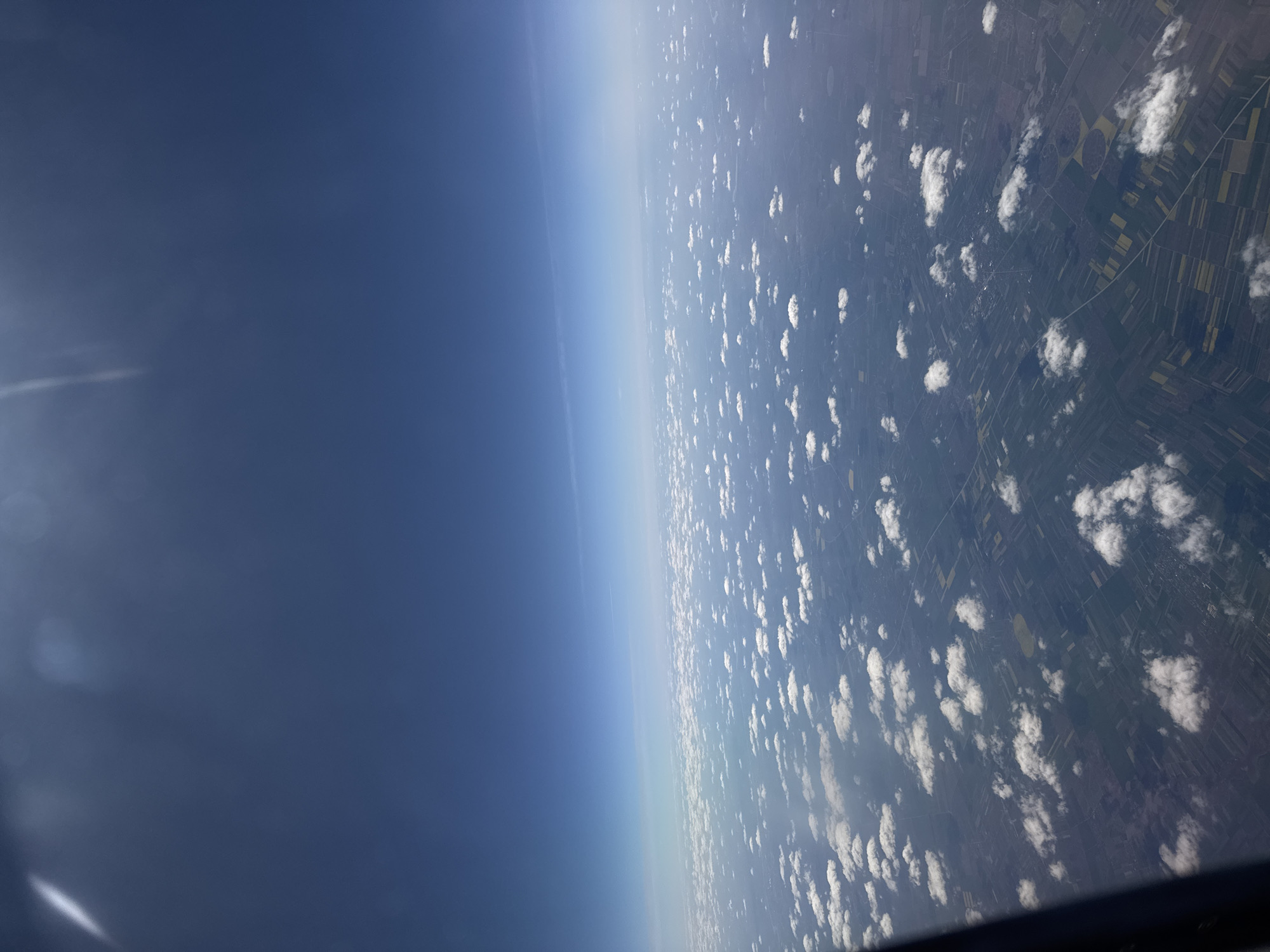 The image displays an aerial view of a landscape and sky, captured from a high altitude, likely from an aircraft. The photograph is oriented with a significant leftward tilt.
The left half of the frame is dominated by a vast expanse of clear blue sky, which darkens to a deep blue at the top-left and lightens to a pale, hazy blue towards the horizon line. This horizon cuts diagonally across the image from the mid-left to the mid-right.
The right half of the image reveals the Earth's surface, covered by scattered white cumulus clouds. Below the clouds, the terrain appears as an extensive patchwork of agricultural fields, varying in shades of green, yellow, and brown, suggesting different crops and cultivation patterns. Distinct circular fields, indicative of center-pivot irrigation, are visible among the predominantly rectangular plots. Faint lines, possibly roads or property boundaries, crisscross the cultivated land.
A subtle atmospheric haze is noticeable above the distant land. In the upper-left corner, a faint, elongated streak of white light or lens flare is present. A dark, angular edge, likely a part of an aircraft window frame, is visible along the bottom and bottom-right edges of the image.
The scene depicts a bright, clear daytime with partial cloud cover. There are no people or discernible text within the image. The landscape characteristics are consistent with agricultural regions, such as those found around Sofia, Bulgaria.