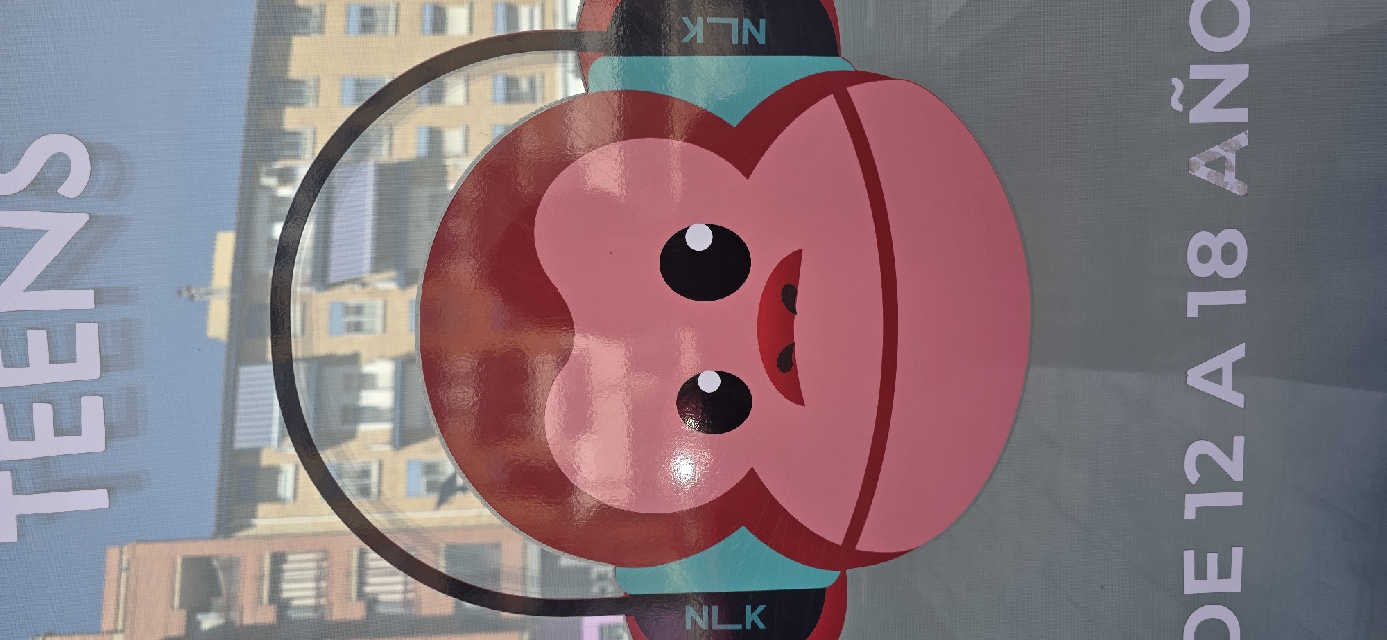 The image displays a portion of a window with a decal featuring a cartoon monkey's face wearing headphones. Above the monkey, the word "TEENS" is visible in large, white, sans-serif lettering. The monkey has large, black eyes with white pupils, a small red mouth, and two dark brown nostrils. Its head is pink, and it is wearing headphones with teal earcups. The text "NLK" appears vertically on the teal earcups. Below the monkey's head, the text "DE 12 A 18 AÑO" is visible in white lettering. The background through the window shows buildings with balconies and windows, suggesting an urban environment. The lighting indicates it is daytime. The scene appears to be a storefront or public display.