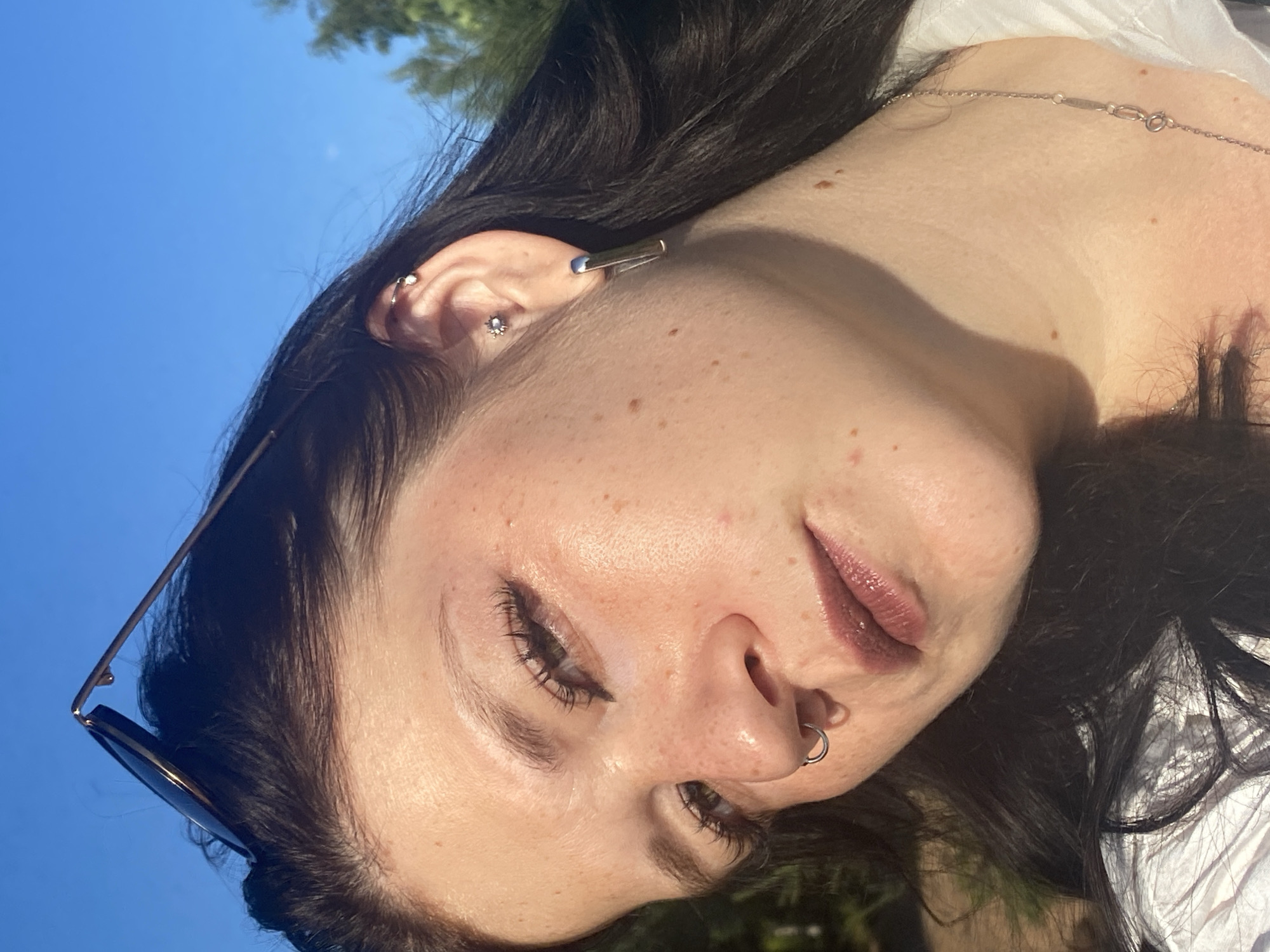 The image is a close-up, angled selfie of a young woman outdoors against a bright blue sky and out-of-focus greenery. She has dark hair, freckles, and is wearing makeup including eyeliner and lipstick. She has multiple piercings, including a septum ring, a cartilage piercing with a small gem, and a lobe piercing with a rectangular earring. She is also wearing sunglasses perched on her head and a delicate silver necklace. The lighting suggests it is daytime, with sunlight illuminating her face from the side. There is no indication of the city of Kostovaty, Russia.