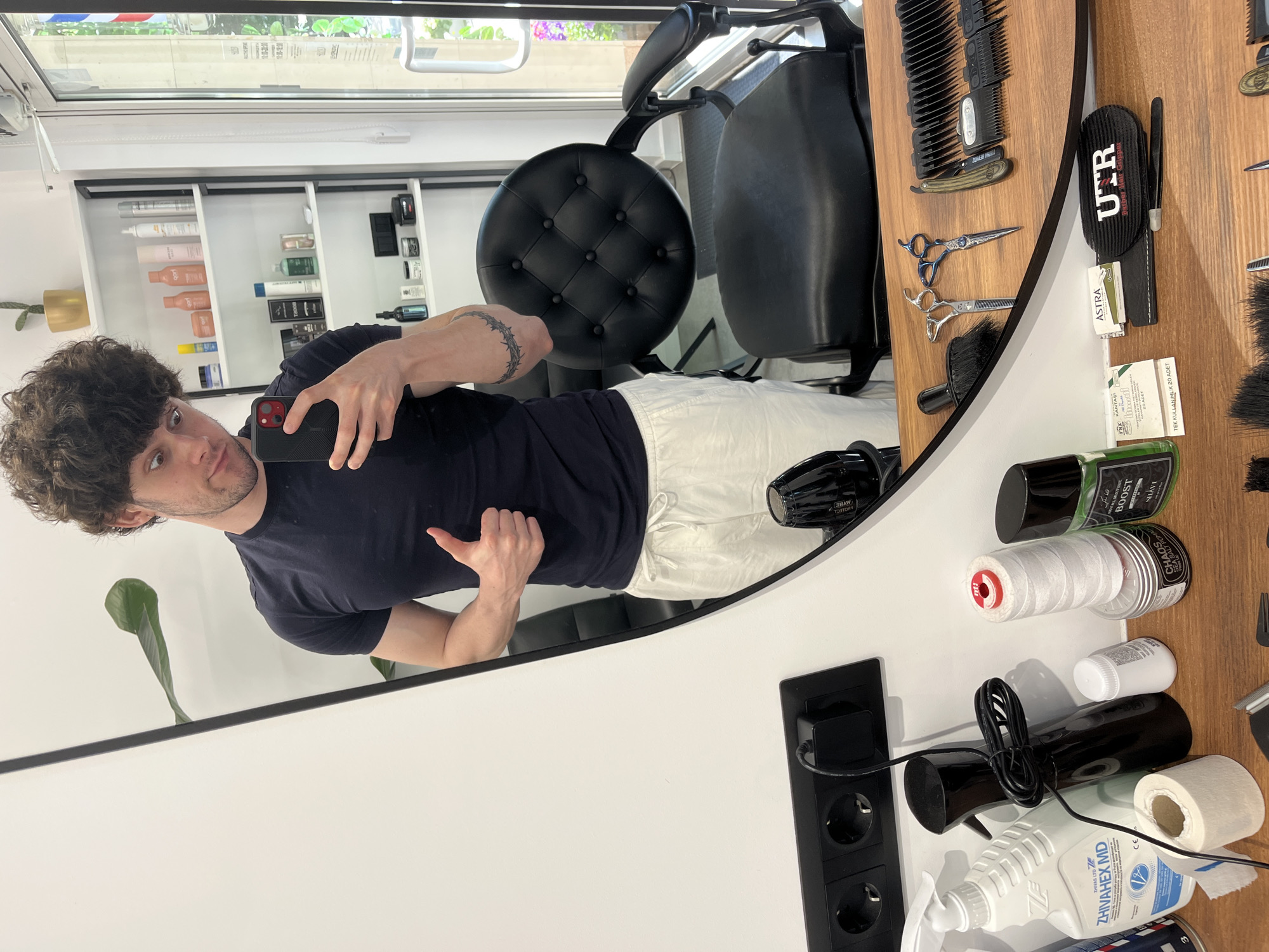 An adult male with dark, curly hair, wearing a dark blue t-shirt and white drawstring pants, is captured in a mirror selfie within a barbershop in Sofia, Bulgaria. He is holding a smartphone with a red case in his right hand, reflecting his image. His left arm is bent, and he is giving a thumbs-up gesture. A tattoo resembling a crown of thorns is visible on his right forearm.
The barbershop interior features a large oval mirror reflecting the man and the environment. Behind the man's reflection, a white shelving unit is visible, holding various hair care products in different bottles and containers, alongside a small cactus in a golden pot. To the far right, a glass door or window reflects outdoor greenery and a faint glimpse of a red, white, and blue barber pole stripe. A black tufted leather barber chair is prominent behind the man's lower reflection.
In the foreground, on a wooden surface below the mirror, various barbershop supplies and products are arranged. These include a white spray bottle labeled "ZHIVAHEX MD," a black spray bottle with a coiled black cord, a roll of white paper tape, and a small white bottle. Also present are a large spool of white thread with a red cap, a clear container of "CHAOS SEA SALT POMADE," a green bottle labeled "ROYAL MANTRA BOOST," and a small package of "KANTASI TNX LINDWOOD" single-use items, along with a small box labeled "ASTRA." A black case marked "UTR BARBER HAIR GRIPPER" and a pair of tweezers are also visible. Professional barber tools, including two pairs of scissors (one with blue handles), a straight razor, several clipper comb attachments, and a black brush, are meticulously laid out on the surface. A black triple electrical outlet is mounted on the white wall below the mirror, with one port occupied by a power adapter.