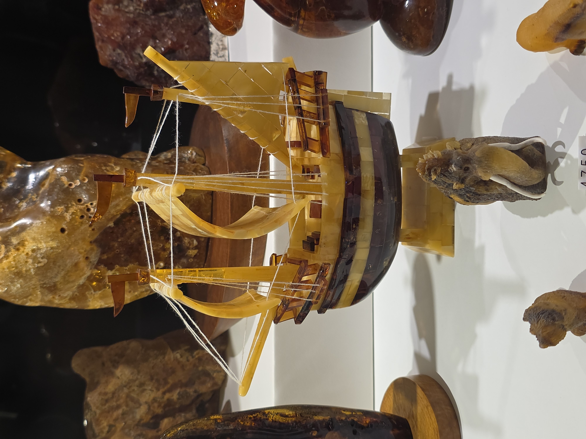 The image displays a handcrafted model of a sailing ship made of amber, exhibited in what is likely a museum or display case. The ship features amber sails, masts with string rigging, and a hull constructed from alternating bands of light and dark amber. It is displayed on a shelf with other amber objects, including what appears to be an amber carving of an animal. The scene appears to be indoors, possibly in a museum in Warsaw, Poland, a location known for its amber craftwork and museums.