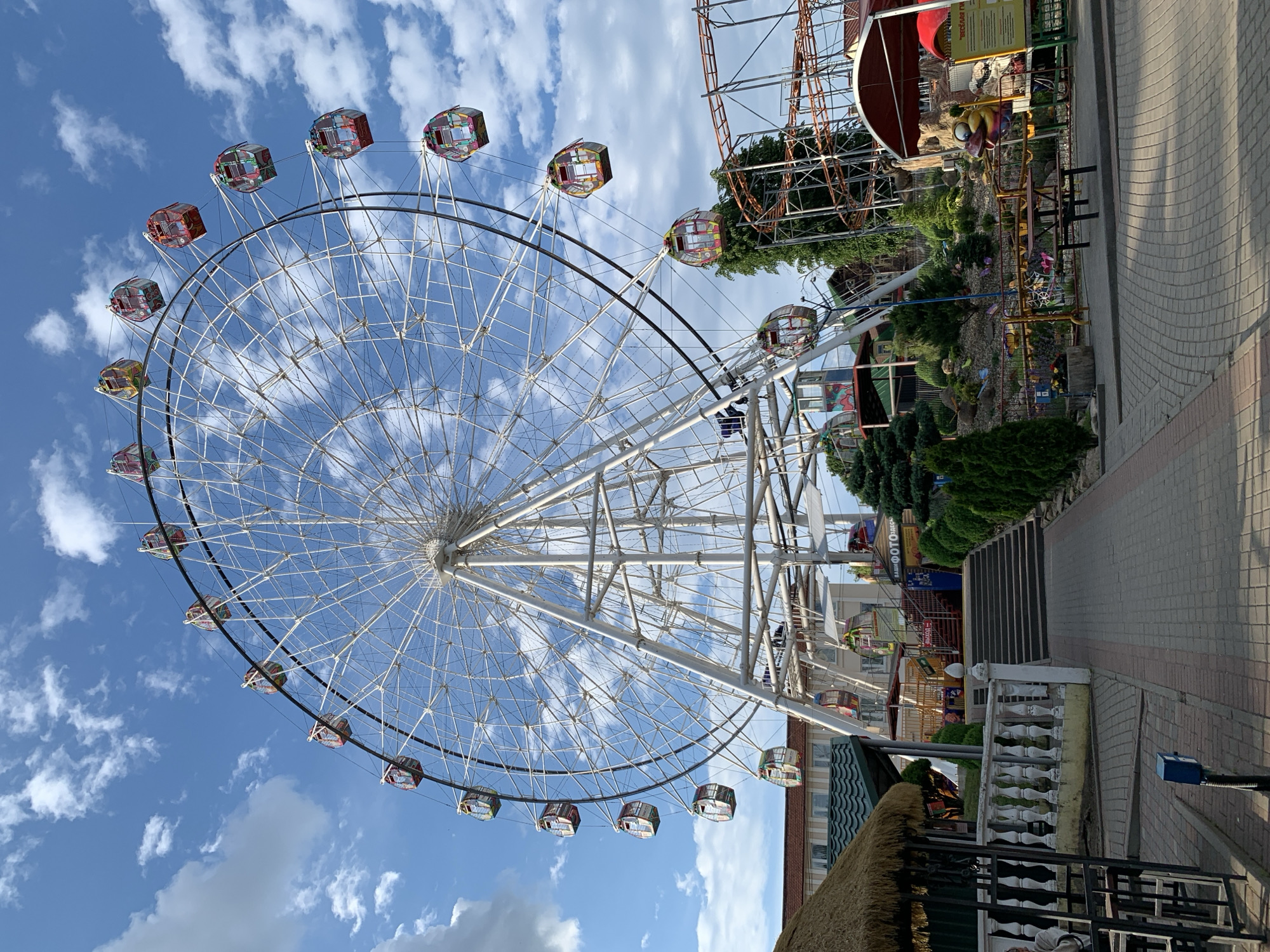 This image depicts an amusement park in Hrodna, Belarus, under a blue sky with scattered white cumulus clouds. A large white Ferris wheel is centrally positioned, its lattice structure and spokes supporting numerous enclosed cabins that display various colors including red, yellow, blue, and green elements. To the right of the Ferris wheel, sections of an orange-brown roller coaster track are visible, supported by metal framework. In the upper left background, a multi-story building with a red-tiled roof and light-colored walls is partially visible. The foreground features a paved area constructed from interlocked pavers, with a white balustrade on the left leading to a straw-thatched gazebo. A set of concrete stairs ascends towards the Ferris wheel, flanked by signs displaying Cyrillic text, including "ФОТО" (PHOTO) and "ВЫХОД" (EXIT). On the right foreground, a landscaped section contains small evergreen trees, decorative rocks, and picnic tables. A red-canopied structure is situated on the far right, near a sign with partial Cyrillic text "ВЕСЁЛАЯ" (MERRY/FUN) and a yellow cartoon character figure. One person is faintly visible seated under the gazebo on the far left. No other prominent human activity or interactions are discernible.