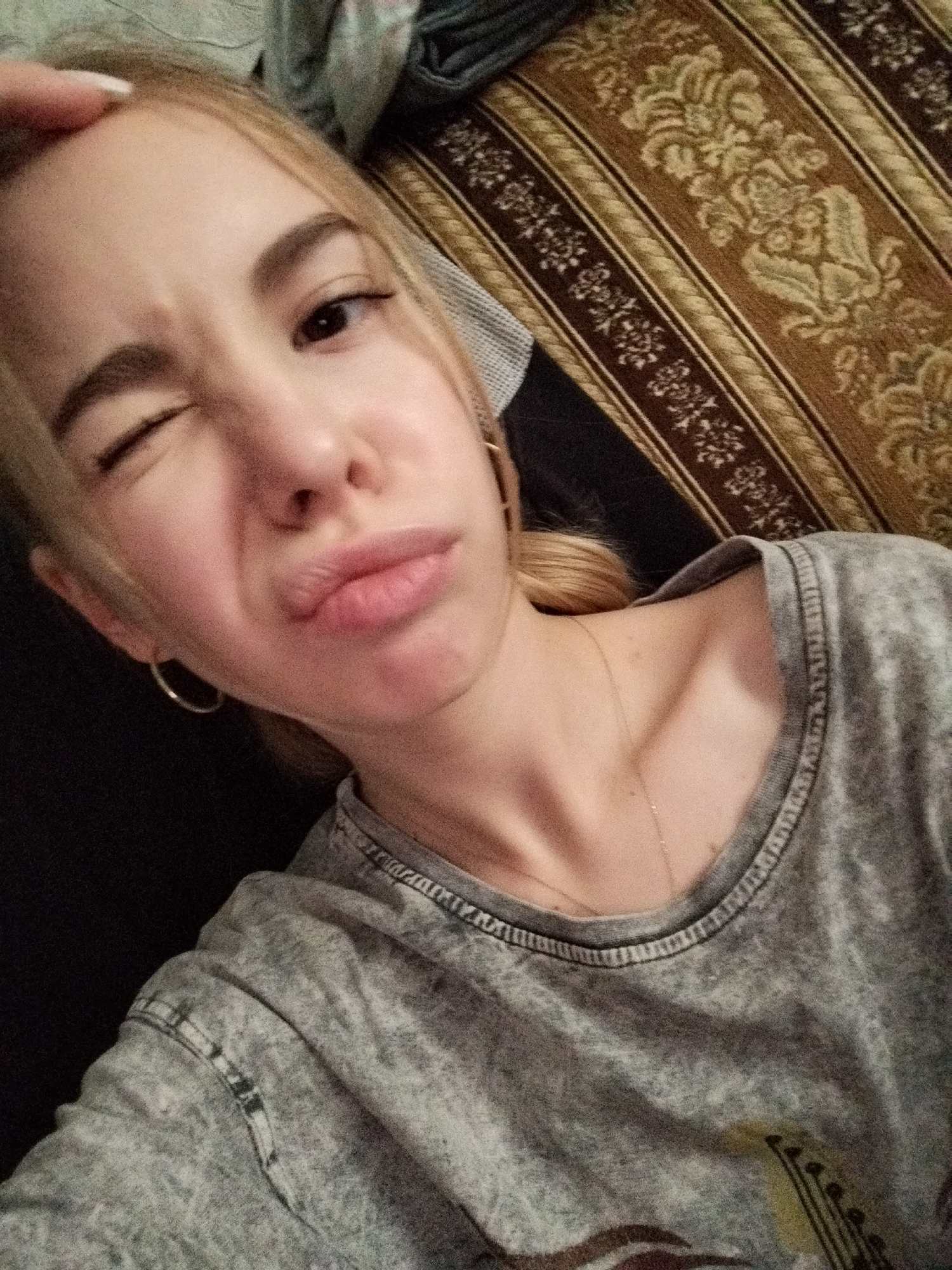 A close-up, overhead shot shows a young blonde woman lying on her side on a couch. She is looking up at the camera with her mouth pursed and one eye winking. The woman is wearing a gray t-shirt and a thin silver necklace. Her left ear is adorned with a small hoop earring. The couch has a brown and gold patterned fabric. The lighting is dim, and the image is slightly blurry. The background is dark and out of focus.