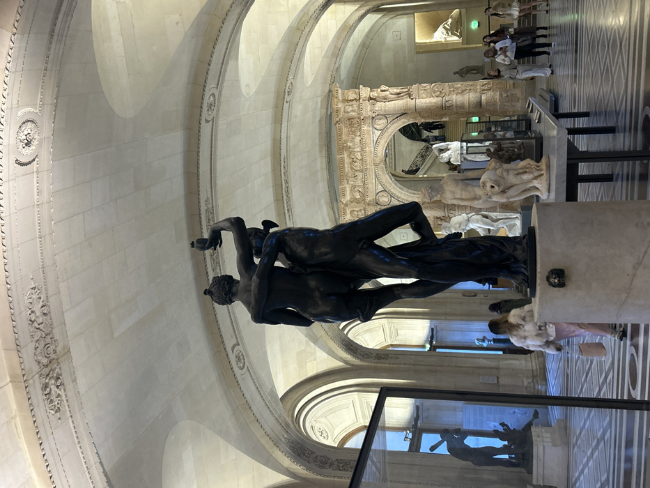 An indoor scene reveals a museum setting with marble floors and arched ceilings. Sculptures on pedestals fill the space, including a dark bronze sculpture depicting two entwined figures. In the background, other sculptures, including an ornate stone archway, are visible. A group of people stands further back, observing the exhibits. Reflections appear in a glass case, mirroring some of the sculptures. The scene appears to be at the Louvre Museum in Paris.