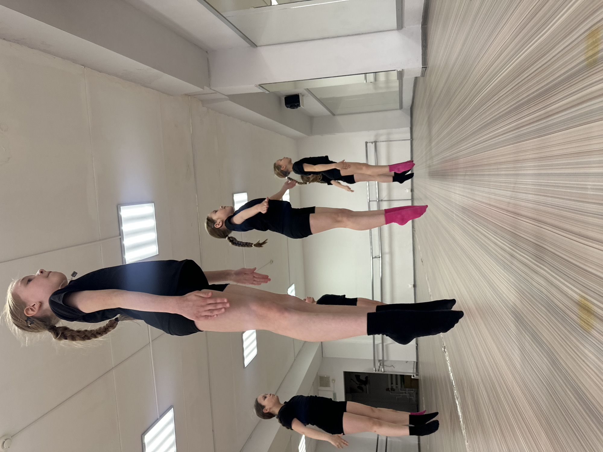 This image captures a group of young females engaged in a dance or gymnastics exercise within a studio setting. The subjects are lying on their backs on the floor, holding a rigid, straight-body pose that appears to be elevated slightly off the ground, with their arms extended alongside their bodies.
They are dressed in dark, form-fitting athletic wear, likely black leotards or matching tops and shorts. Several individuals are wearing brightly colored socks; for example, the prominent figure in the foreground wears vibrant pink split-toe socks, while others have black socks, some also appearing to be split-toe. Their hair is neatly tied back, with at least one having a long braid.
The setting is characterized by a large mirrored wall on the left, which reflects the subjects and the white ceiling with fluorescent lighting. The floor features a distinctive pattern of light-colored, thin parallel lines, creating a sense of depth and texture. The overall impression is one of focused concentration during a practice session. There is no visible text in the image. The scene takes place indoors, with artificial lighting, in what appears to be a dedicated training facility in Votkinsk, Russia.