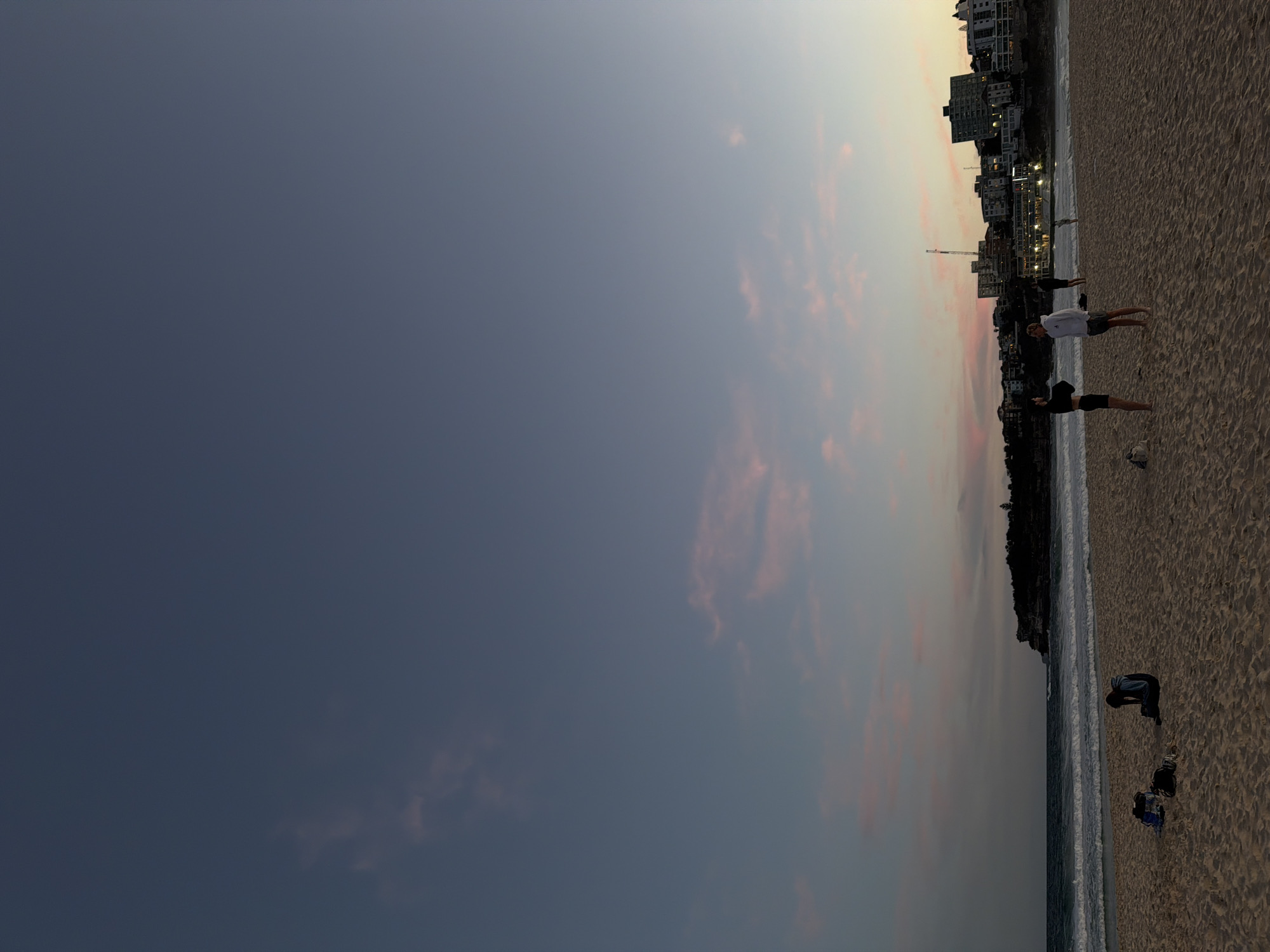 The image depicts Bondi Beach in Sydney, Australia at dusk. The sky is a gradient of deep blue at the top transitioning to lighter hues of blue and pink near the horizon, with scattered wispy clouds. The ocean is visible in the mid-ground, with gentle waves breaking on the shore. The beach itself is sandy and mostly empty, with a few scattered items like bags.
In the foreground, a person is sitting on the sand, facing away from the camera. Further into the scene, near the water's edge, a group of four people are standing and interacting. Two of them appear to be conversing, while the other two are standing nearby. In the background, buildings line the coast, with some lights illuminating the structures. A crane is also visible among the buildings. The overall scene conveys a sense of a quiet evening at a popular beach location.