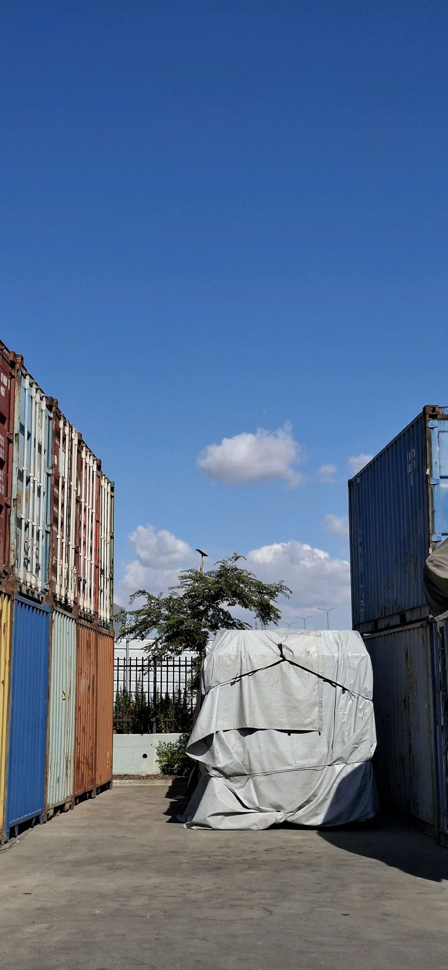 An outdoor shot captures a stack of shipping containers on a concrete surface, bathed in sunlight under a bright blue sky. On the left, several brightly colored shipping containers are stacked, showcasing shades of red, white, blue, and orange. To the right, a larger, dark blue shipping container is visible, partially obscured by a covered object in the foreground. This object is shrouded in a light gray tarp, tightly secured with ropes, creating an irregular, lumpy shape. Behind the covered object, a small tree with dark green foliage stands beneath a pole with a solar panel. A black metal fence borders the area behind the tree, with dense green foliage visible beyond it. The sky is clear except for a few scattered white clouds. The scene appears to be a port or industrial area, possibly in the city of Kiryat Gat, Israel, given the context. No people are present in the image.