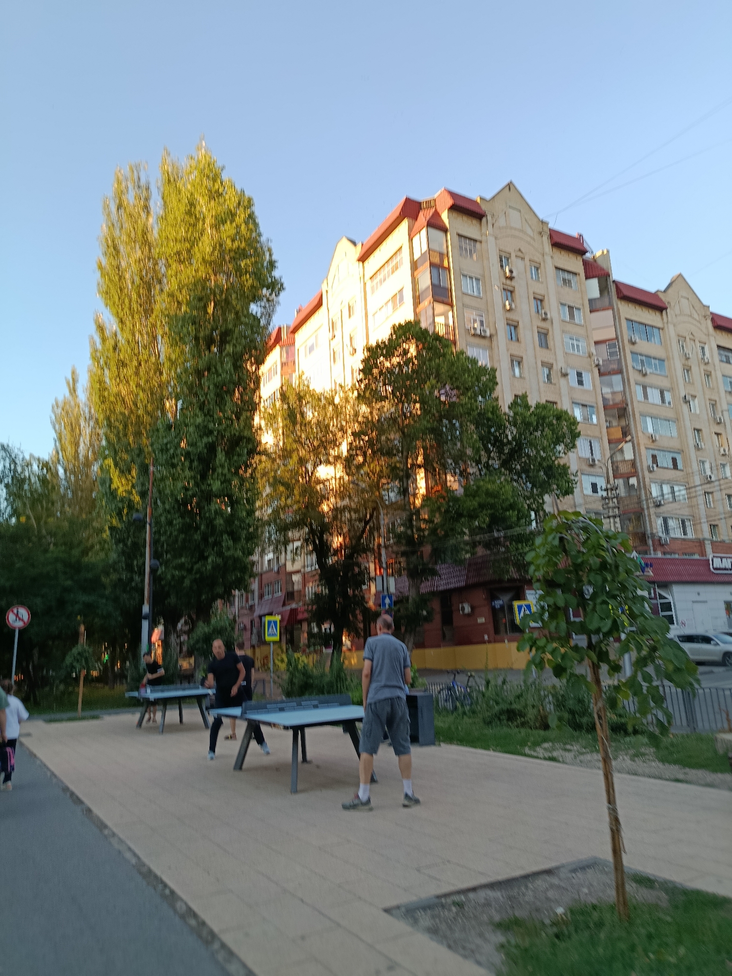 The image depicts an outdoor scene with a building and people engaged in recreational activities. The building, a multi-story structure with a red roof and numerous windows, appears to be residential or mixed-use. In front of the building, there are several outdoor ping pong tables, and individuals are seen playing or standing near them. The environment suggests a public space or a recreational area within a city. The presence of trees and a clear sky indicates a daytime setting, likely during warmer weather. Based on the architectural style of the building and the overall urban setting, this scene is consistent with an area in Saratov, Russia.