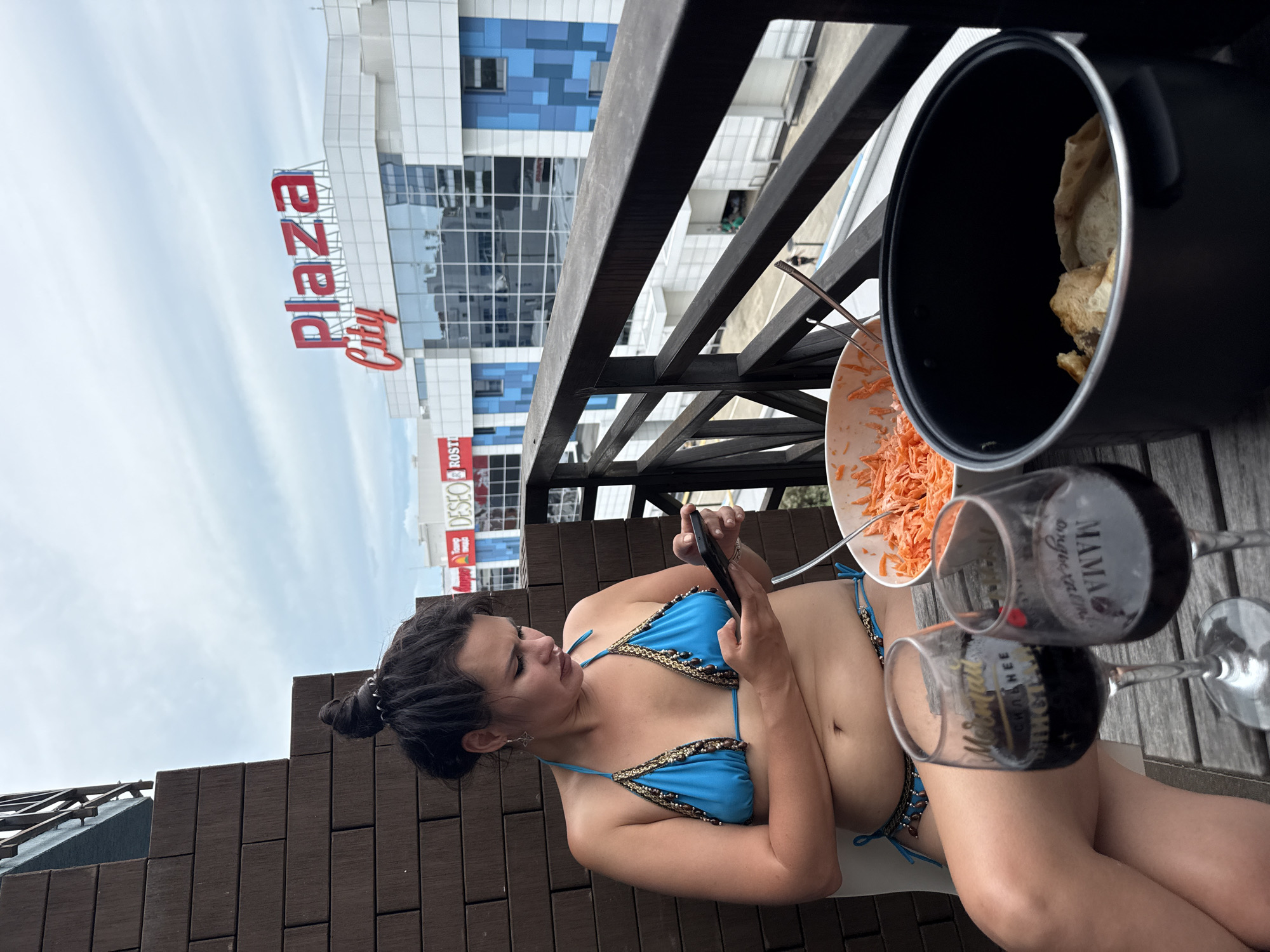 A woman in a blue bikini is sitting on a balcony with a smartphone in her hands. She is looking down at the phone. In front of her on a table are two glasses of red wine, a bowl of shredded carrots, and a pot with bread. In the background, a large building with "Plaza City" signage is visible, indicating the location is in Sochi, Russia. The sky is cloudy.