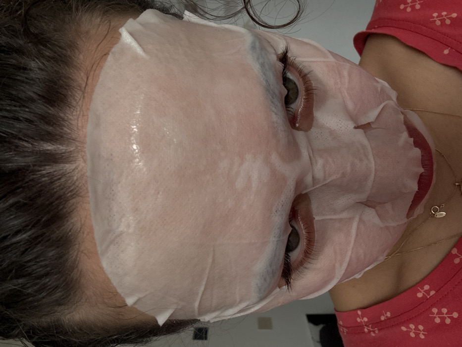 The image displays a close-up, downward-angled view of a person wearing a translucent white sheet face mask. The mask covers the forehead, cheeks, nose, and chin, revealing the eyes and mouth through cutouts. The person's dark hair is visible above the mask and partially on the right side of the head. Dark green eyes with prominent eyelashes are visible, along with dark eyebrows just above the mask's eye openings. Red-tinted lips are visible beneath the mask's mouth opening. The person is wearing a red-orange shirt with a repeating white leaf-like pattern on the left upper chest area, and a thin gold-colored necklace with small pendants is visible. The background is out of focus, showing indistinct light-colored walls with faint rectangular and square shapes. The scene takes place in the city of Milan, Italy.