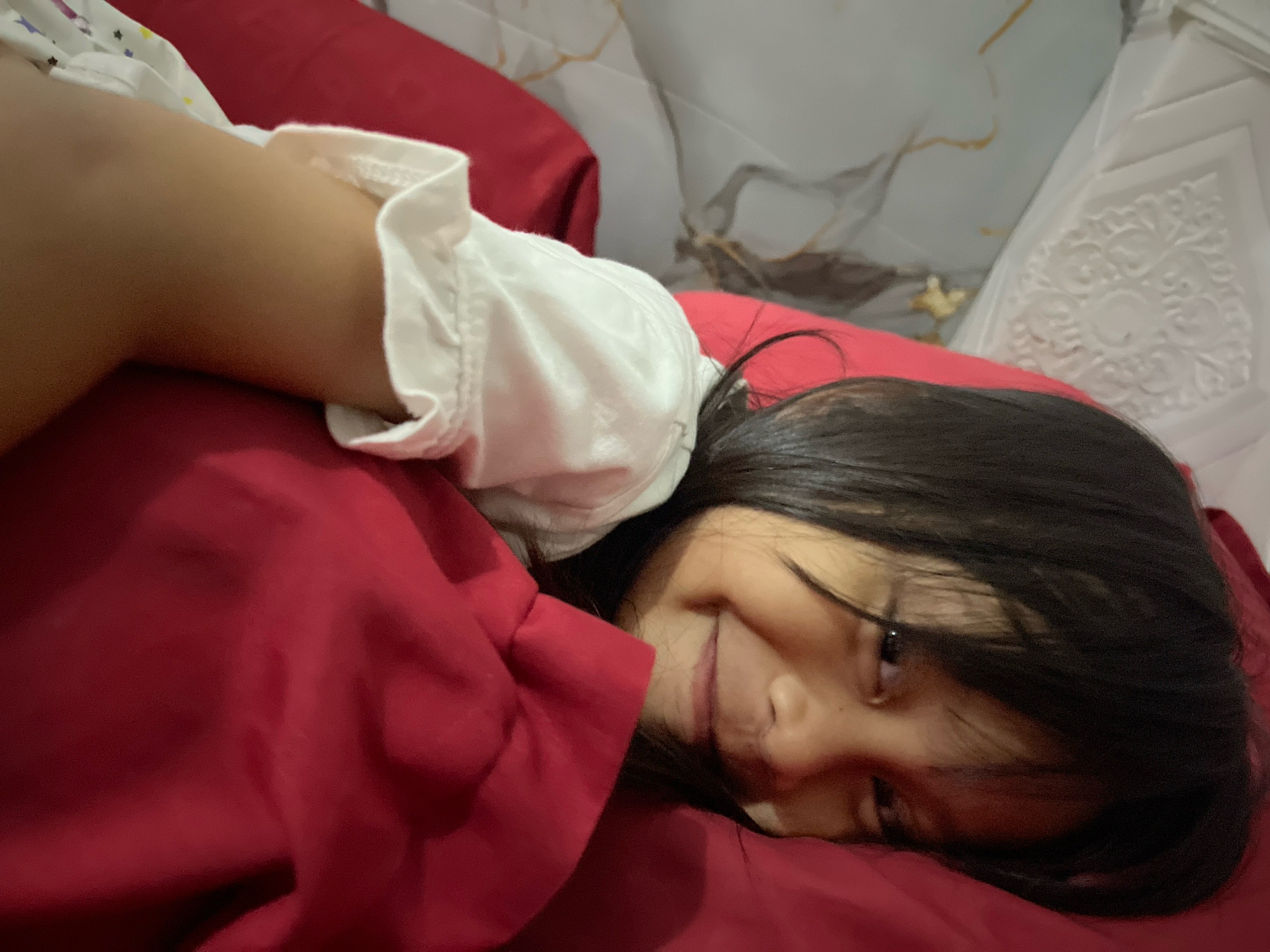 A child lies on a red surface, possibly a bed, with their head resting on a pillow. The child's left arm, wearing a white shirt with a patterned sleeve, is draped over their chest. The child's face is turned towards the camera, with their eyes closed and a slight smile. The background consists of a white, textured surface and a red pillow. The setting appears to be indoors, likely a bedroom, within Kecamatan Pati, Indonesia.