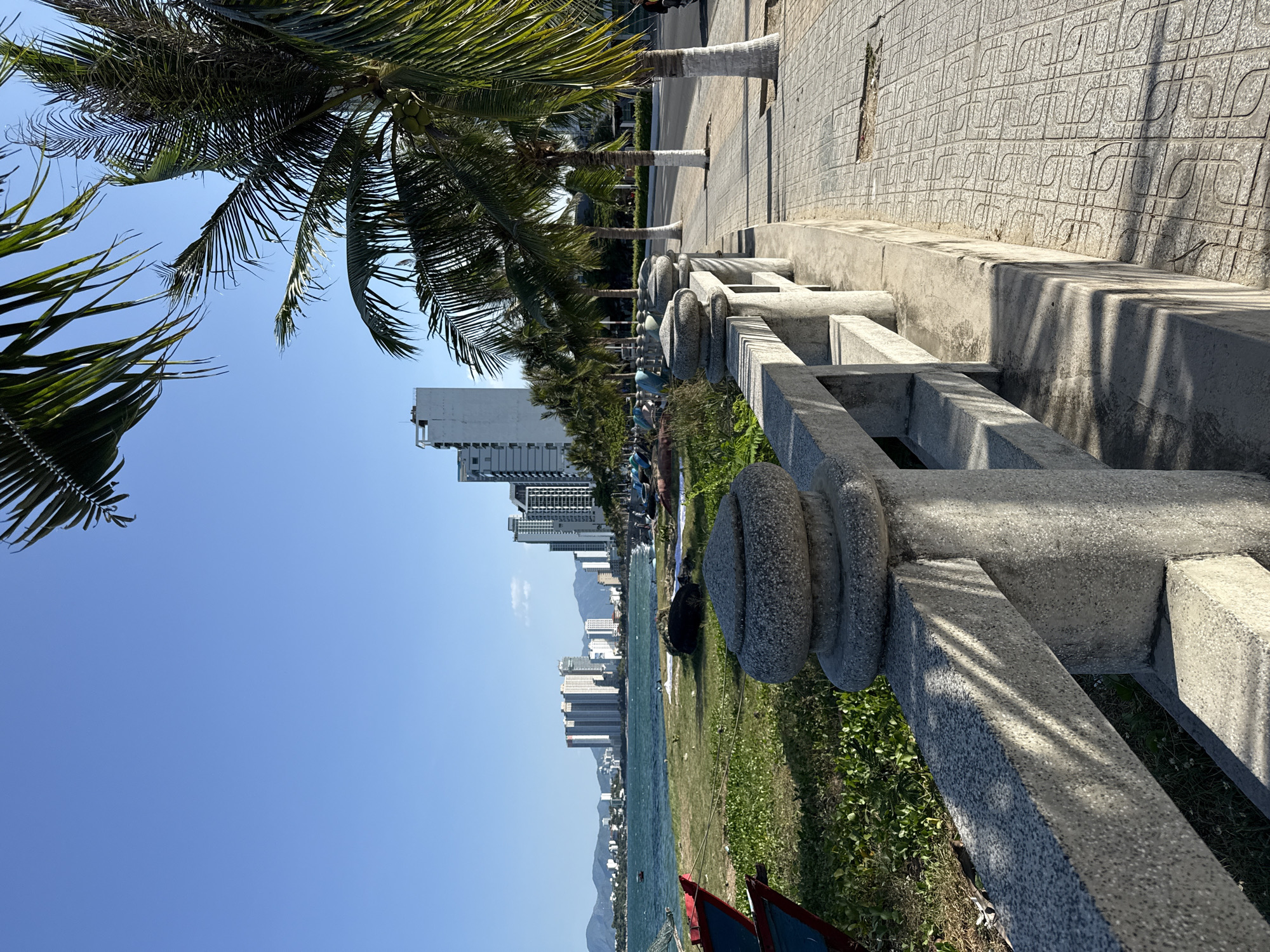 A scenic view of the coastline in Bac Nha Trang Ward, Vietnam, is depicted. The foreground features a grey concrete railing with decorative, bulbous stone posts, casting shadows from palm fronds overhead onto the walkway. The walkway itself is paved with textured square tiles arranged in a geometric pattern. Beyond the railing, a strip of green grass and foliage separates the walkway from the sea. Several fishing boats are partially visible on the grassy area. The sea is a vibrant turquoise, with waves lapping against a distant shore. In the background, a city skyline is dominated by numerous tall, modern buildings, stretching across the horizon. Mountainous terrain forms the backdrop to the city. Coconut palm trees with visible coconuts line the road adjacent to the walkway, their fronds reaching towards the clear blue sky. The sun is high, indicating daytime.