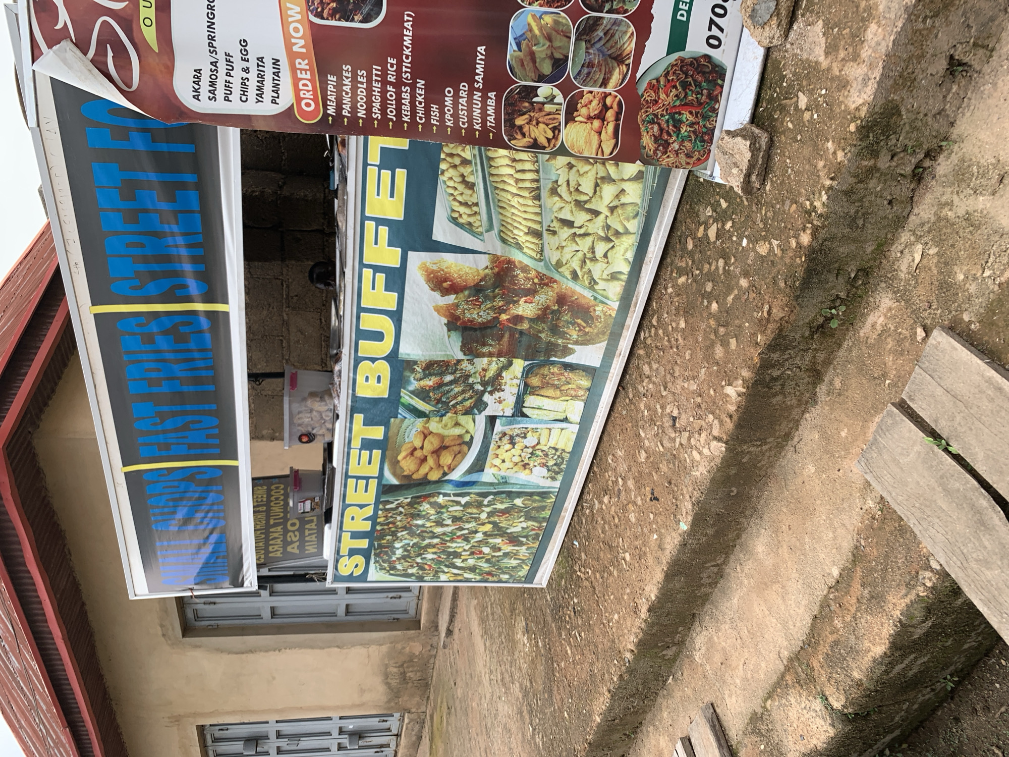 This media file depicts a street food stall in Jos, Nigeria. The stall features a large, colorful sign advertising "STREET BUFFET" with images of various dishes, including fried fish, fried plantains, and noodles. Another sign, partially obscured, reads "FAST FRIES" in large blue letters. Text on the "STREET BUFFET" sign lists items like Akara, Samosa, Puff Puff, Chips & Egg, Yamarita, Plantain, Meatpie, Pancakes, Noodles, Spaghetti, Jollof Rice, Kebabs, Chicken, Fish, Kpomo, Custard, and Kunun Samiya. A phone number, "070", is also visible. The stall is set up outdoors, with a brick wall behind it and a rough, earthy ground in front. The lighting suggests it is daytime, with overcast or diffused light. The overall impression is of a bustling local eatery offering a variety of popular Nigerian street food options.