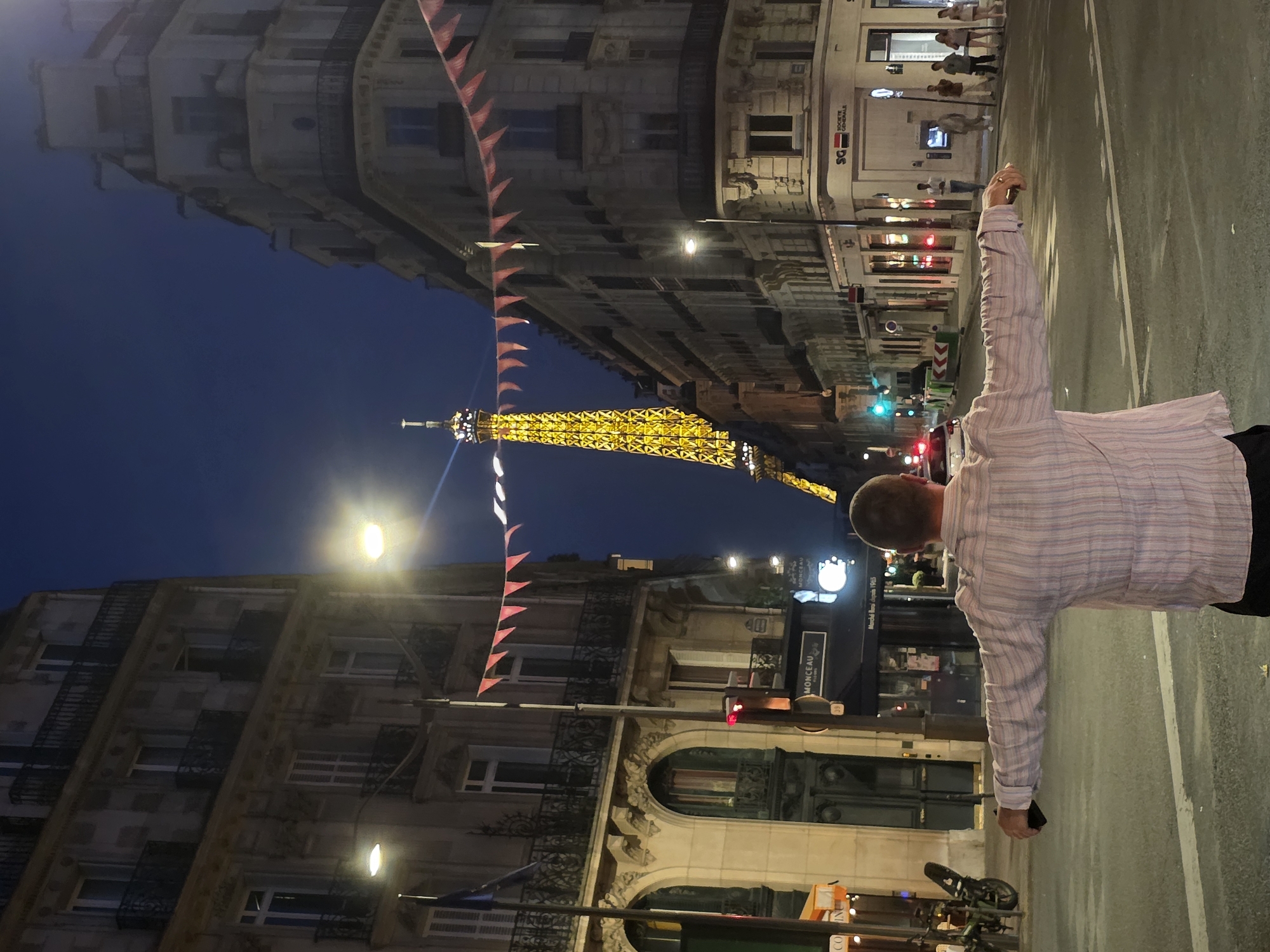 The image depicts a street scene in Paris, France, at dusk. The Eiffel Tower, illuminated in yellow, is visible in the background. A man, facing away from the camera, stands in the middle of the street with his arms outstretched. He is wearing a light-colored, striped shirt and dark pants. The street is lined with buildings, some of which have balconies and ornate architectural details. Streetlights are on, and there are small red and white pennant flags strung across the street above the buildings. A few people are visible walking on the sidewalk in the distance. A white car and some traffic cones are also present in the street. The overall impression is a metropolitan evening in Paris, with a tourist capturing the iconic landmark.