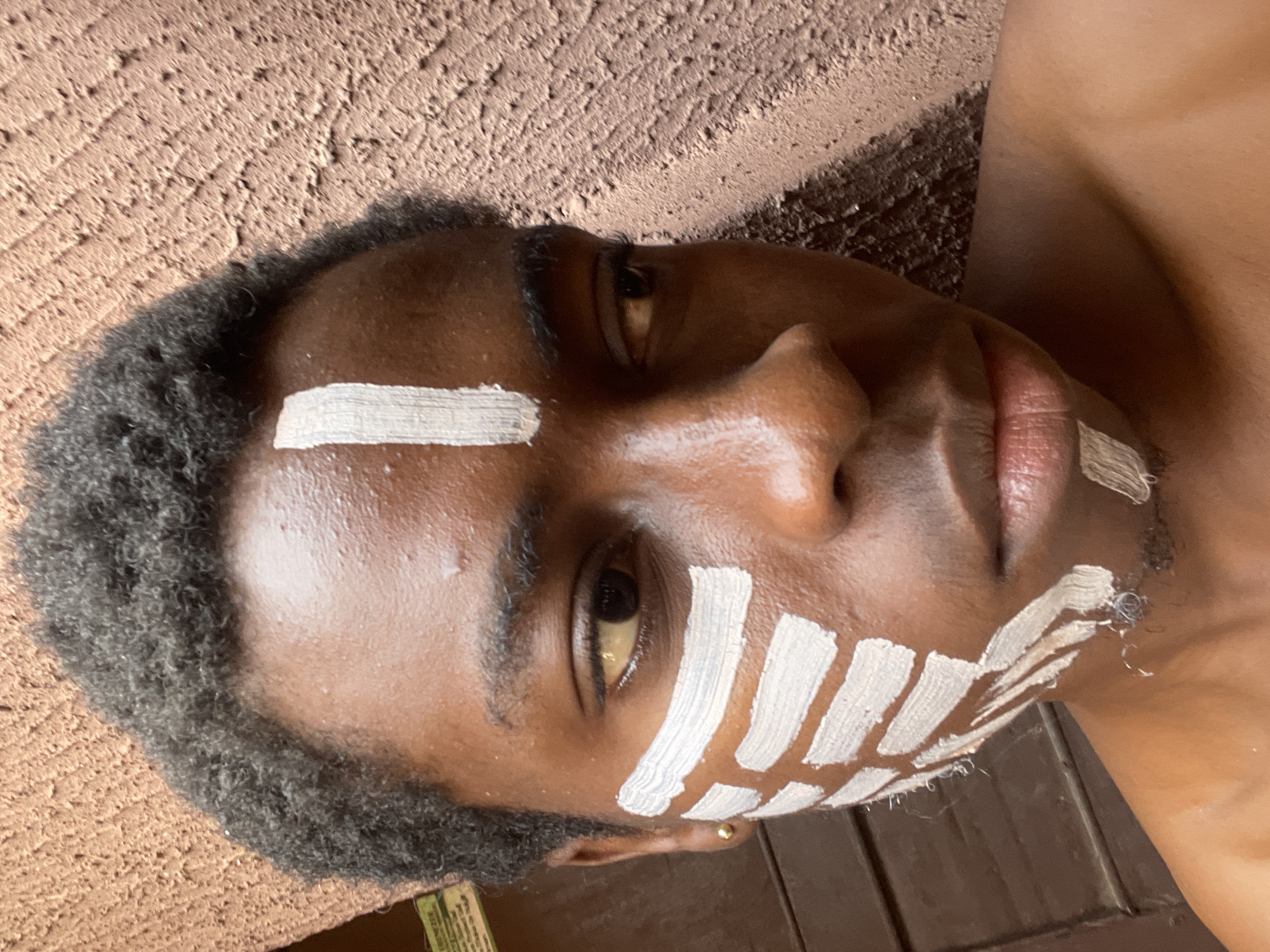 The image displays a close-up portrait of a young person, likely male, with dark skin and short, dark, curly hair. The individual's face is strikingly adorned with white paint markings, applied in thick, horizontal and diagonal stripes across the forehead, beneath the right eye, and on the right cheek and jawline. A small, golden earring is visible in the right earlobe. The person's eyes are open and directed towards the viewer, exhibiting a neutral to slightly serious expression, with the mouth slightly ajar.
The background features a textured, rough-surfaced wall in tones of brownish-orange, suggesting an outdoor or rustic environment. The bright lighting indicates that the photo was taken during the daytime. The person appears to be lying down or leaning against this textured surface, and the image is tilted at an angle. The cultural face paint suggests a ceremonial, traditional, or artistic event taking place in Onitsha, Nigeria, as provided by the context. No legible text is visible within the image frame.