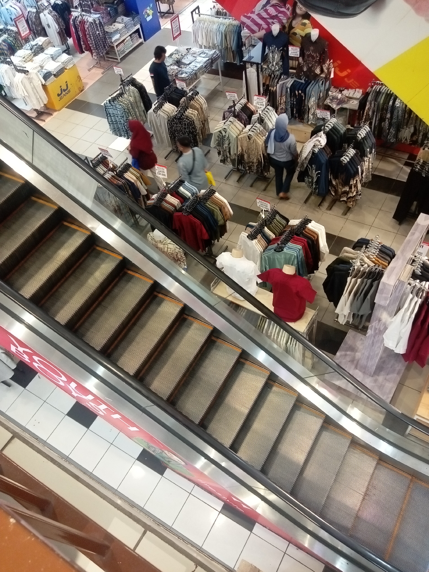 The image depicts an interior view of a department store, likely a clothing section, from an elevated perspective looking down at an escalator. The escalator, with its metallic steps and handrails, dominates the left side of the frame, descending into the lower level of the store.
On the right side of the escalator, various clothing racks filled with garments are visible. The clothes appear to be predominantly shirts and tops, displayed in a variety of colors and patterns, some on hangers and others folded on shelves or mannequins. Several price tags with Indonesian Rupiah denominations are also discernable, such as "Rp154.000" and "Rp174.000."
A few individuals are present in the scene. On the escalator itself, a person wearing a blue headscarf and a grey top is moving upwards. Below the escalator, a person in a dark shirt is standing near a clothing display. Further back, a person in a red headscarf is seen walking amongst the clothing racks. Another individual in a blue hijab is observed looking at garments. The floor is tiled with a pattern of white and black squares. In the background, more clothing displays and store fixtures are visible, including what appears to be a large banner with colorful graphics.
Based on the retail context and the Indonesian currency displayed on the price tags, this scene is likely located in a department store in Batam, Indonesia. The overall impression is of a busy retail environment during shopping hours.