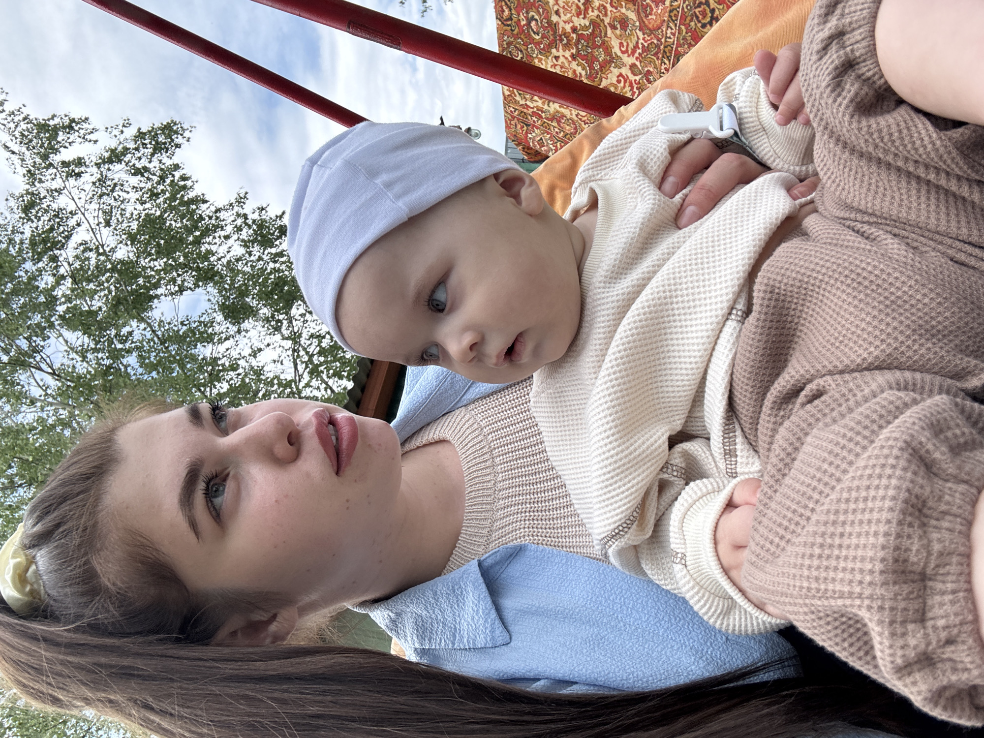 A woman and a baby are the subjects of an outdoor photograph. The woman is lying down, her head towards the bottom of the frame. She has long brown hair partially pulled back with a yellow scrunchie, fair skin, and is wearing a light blue textured shirt. The baby, wearing a white cap, lies on top of her. The baby is wearing a beige waffle-patterned top. A hand is visible on the baby's leg.
Above them is a red structure, likely part of a swing set or outdoor frame. Behind the red structure, there is a decorative rug or blanket. In the background, out of focus, are green trees and a cloudy sky. The photograph is taken from a low angle, looking upwards.
The city is unspecified, only Мещёры, Russia is identified.