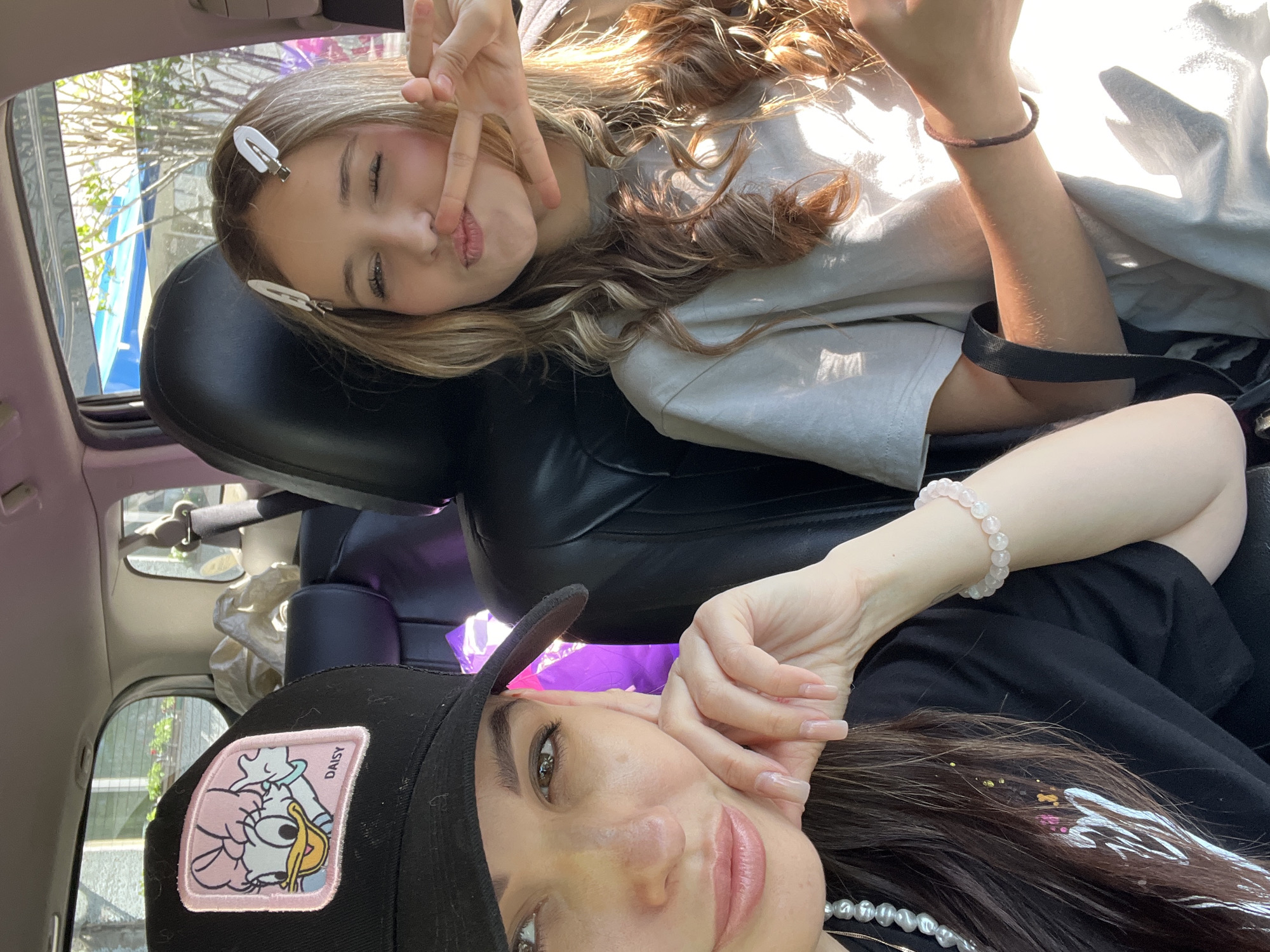 Two individuals are visible inside a vehicle. The upper person has blonde hair, a white barrette, and is making a "peace sign" gesture. The lower person is wearing a black hat with a Daisy Duck patch and has dark hair. The interior of the vehicle includes black leather seats. The environment suggests an outdoor setting, though specific details about the city are not discernable. The actions include posing for a photograph.