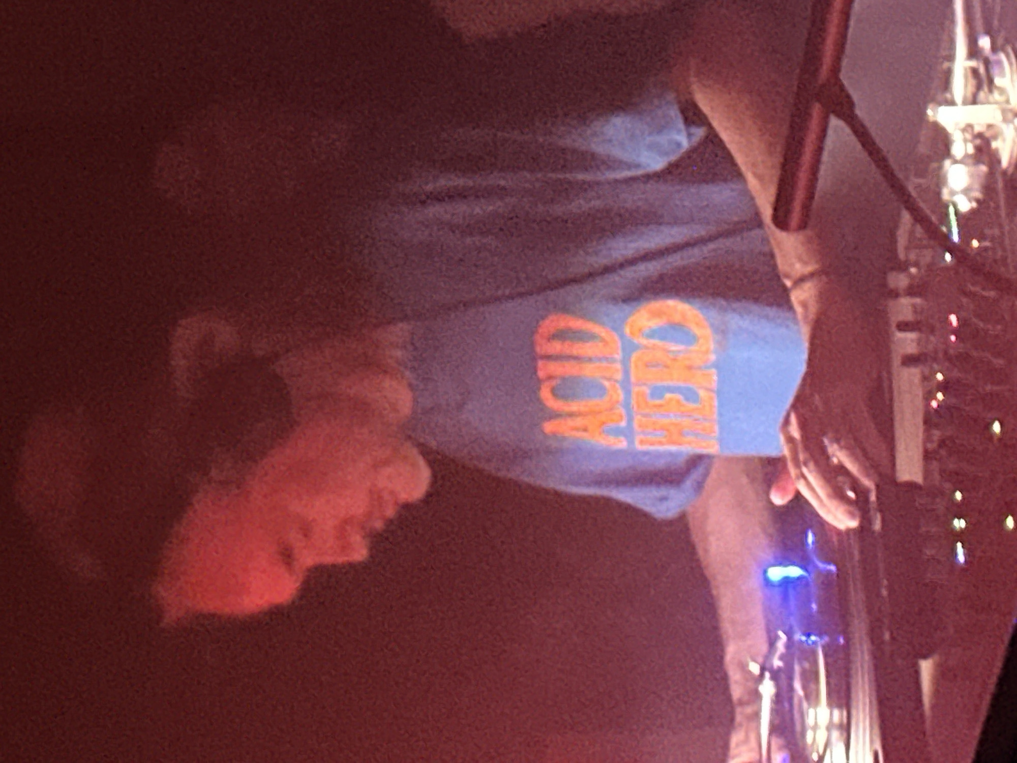 A medium shot reveals a person wearing a blue t-shirt with the words "ACID HERO" printed in orange. The person is facing left with their head visible in the red-lit foreground. Their right hand is visible operating equipment with glowing blue lights. To the right, a stand and other equipment with multiple lights are visible. The location appears to be a performance venue, potentially in Paris, France. There is no definitive information to ensure the city location is Paris, France.