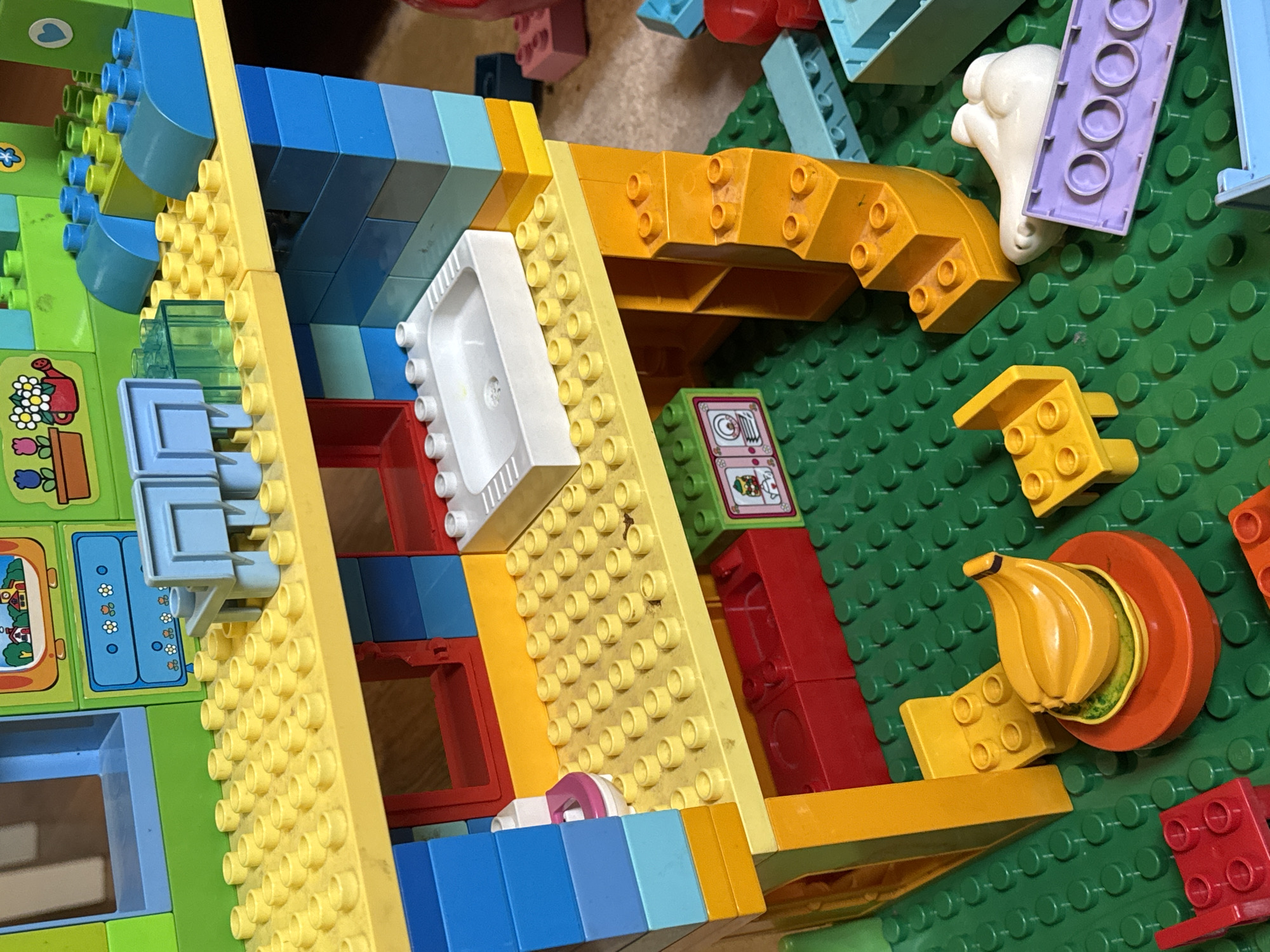 The media file displays a close-up, overhead view of a collection of brightly colored plastic toy bricks, primarily in shades of yellow, blue, red, and green, arranged on a green baseplate. The bricks are consistent with the style of building blocks designed for young children.
Several distinct objects are constructed or represented using these bricks. Prominently featured is a white toilet with a pink lid, positioned on a yellow surface. Adjacent to this is a yellow structure with steps, and further to the right, a cluster of bright yellow banana-shaped bricks sits within an orange circular piece. Other visible elements include a white, cloud-like plastic shape, a purple rectangular brick with circular indentations, and a green and white sticker depicting a cat wearing a party hat and holding balloons. There are also several smaller, single-color bricks and some translucent blue pieces.
The arrangement suggests a partially built structure or a scene created from these toys. The background is a green baseplate with the characteristic circular studs used for connecting toy bricks. The overall impression is one of a children's play area, with the toys meticulously arranged on a play mat or floor. There are no discernible people, animals, or indications of time of day or weather. The location context provided is Zassovskaya, Russia, but this cannot be visually confirmed from the image itself. There is no visible text within the image.