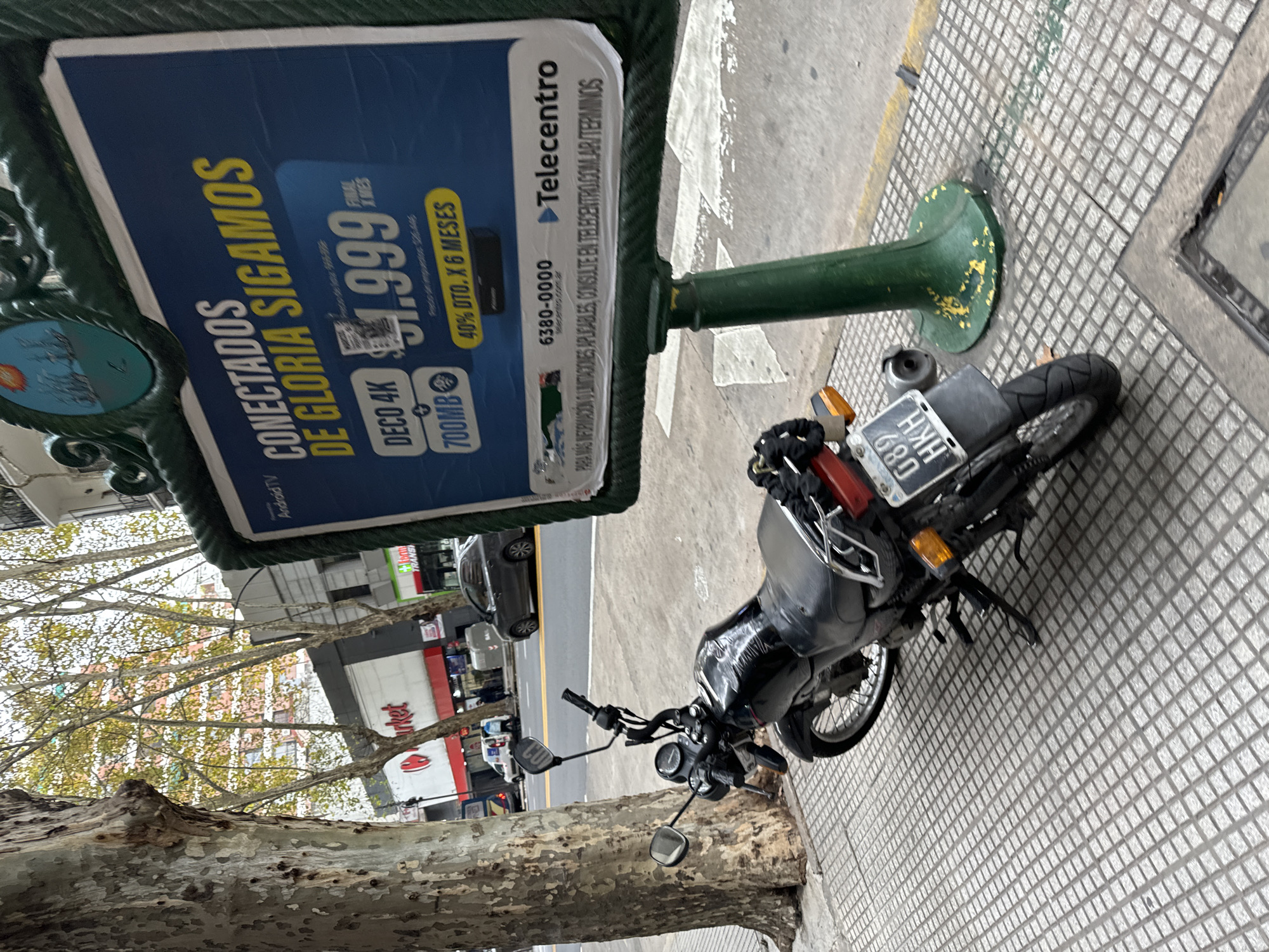The image shows a Telecentro advertising display on a post at street level next to a motorcycle parked on a checkered sidewalk. The sign advertises a Deco 4K product with 700MB for $1,999 in Argentina, featuring the slogan "CONECTADOS DE GLORIA SIGAMOS". A tree trunk is positioned to the left of the sign and motorcycle.
In the background, across the street, is a "Market" store sign. Further down the street, there are a bus and other vehicles. Based on the architecture and environment, the image is likely taken in Buenos Aires, Argentina. The motorcycle's license plate reads "HKH 689."