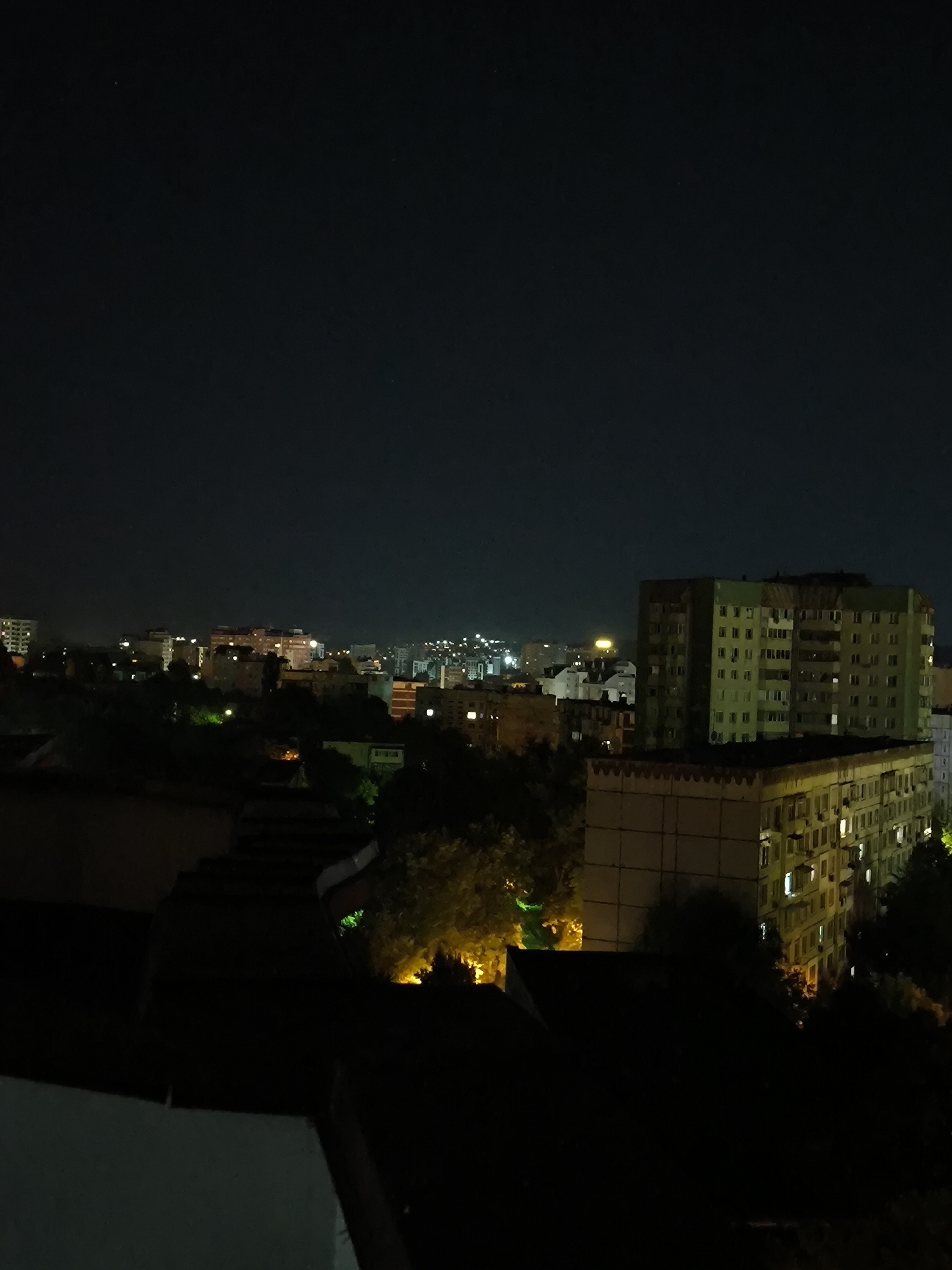 The image is a night-time aerial view of a cityscape, likely Chișinău, Moldova. The dark sky is illuminated by the lights of apartment buildings and streetlights in the distance. In the foreground and middle ground, several multi-story apartment buildings are visible, some with windows lit from within, suggesting occupancy. Trees are scattered throughout the scene, their foliage dark against the night sky, with some illuminated by artificial lights. The architecture appears to be typical of Soviet-era residential blocks. The overall impression is one of a quiet urban night, with the city's infrastructure and residences softly glowing under the darkness.
